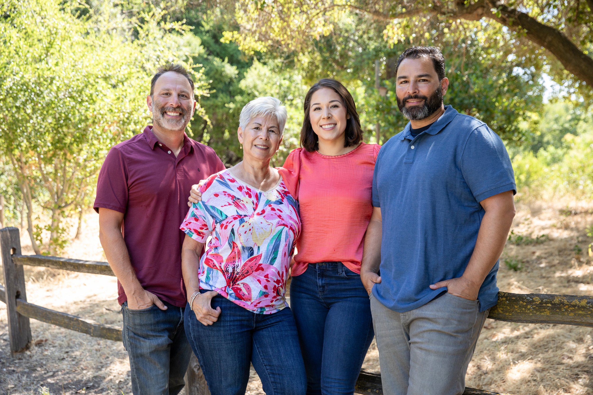 A diverse group of four people smiling outdoors on a sunny day, standing near a wooden fence with trees and greenery in the background.