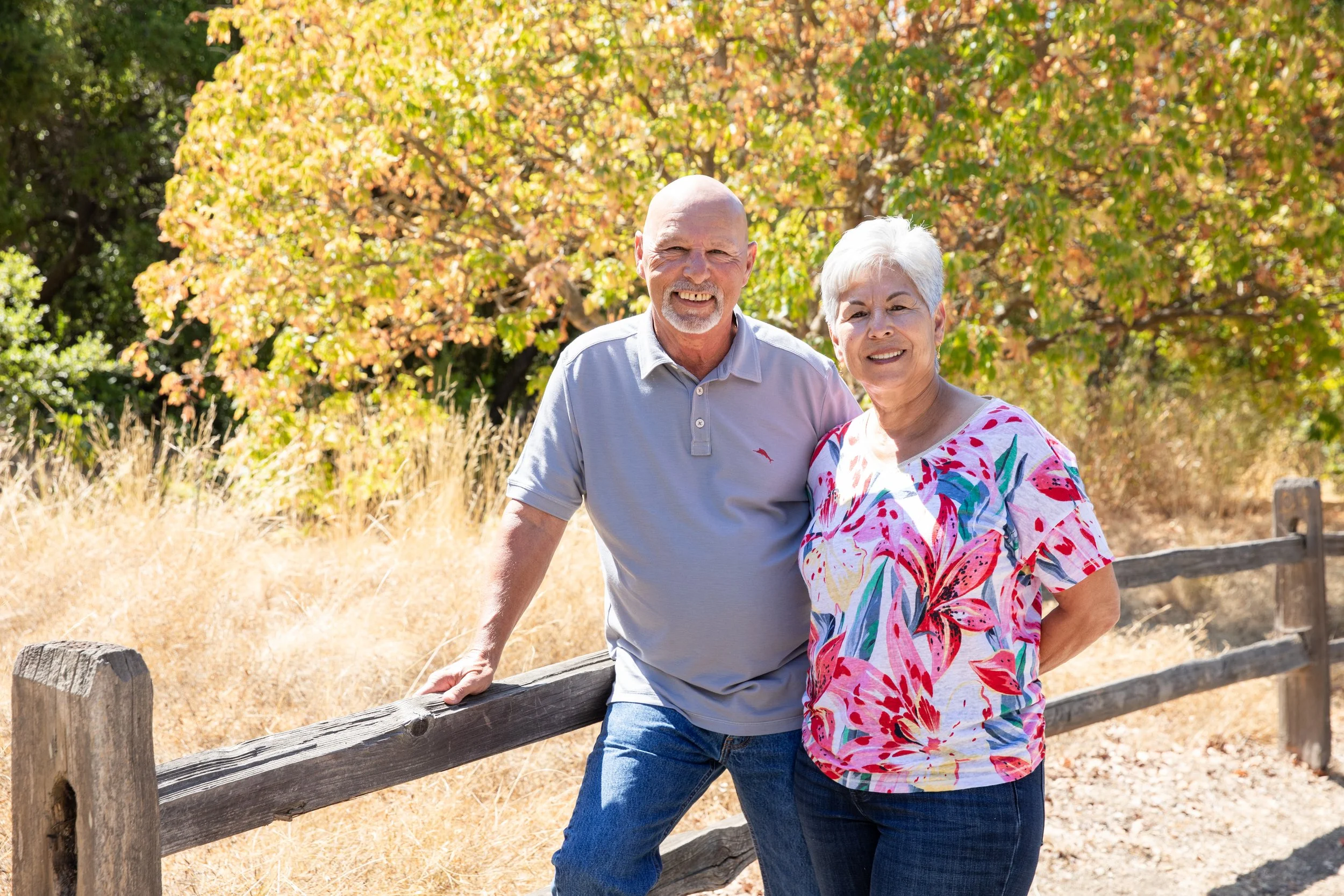 An elderly couple smiling outdoors on a sunny day, standing behind a wooden fence with trees showing fall foliage in the background.