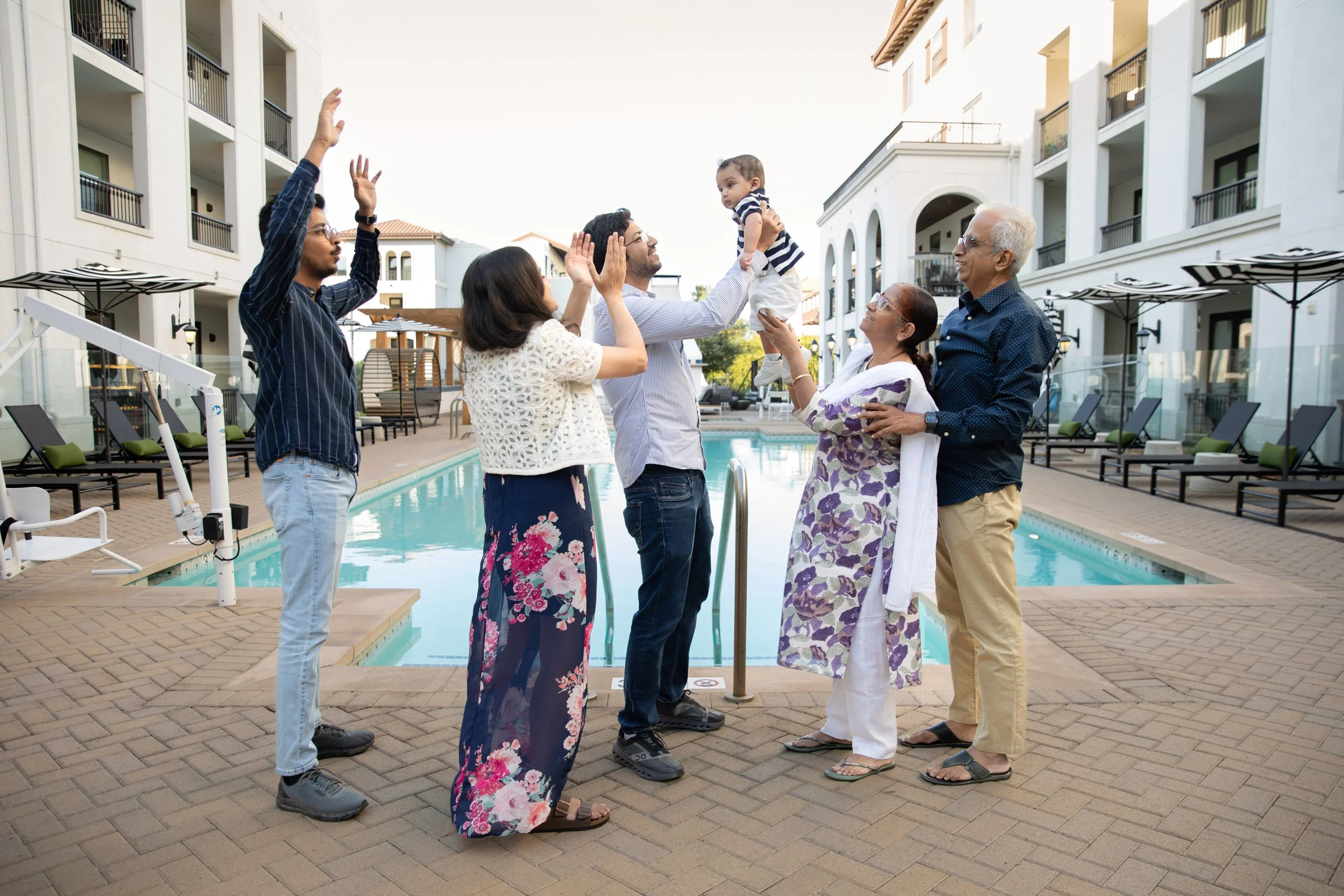 A multigenerational family enjoying time by a swimming pool, with a young child being lifted and celebrated, in an outdoor residential complex.