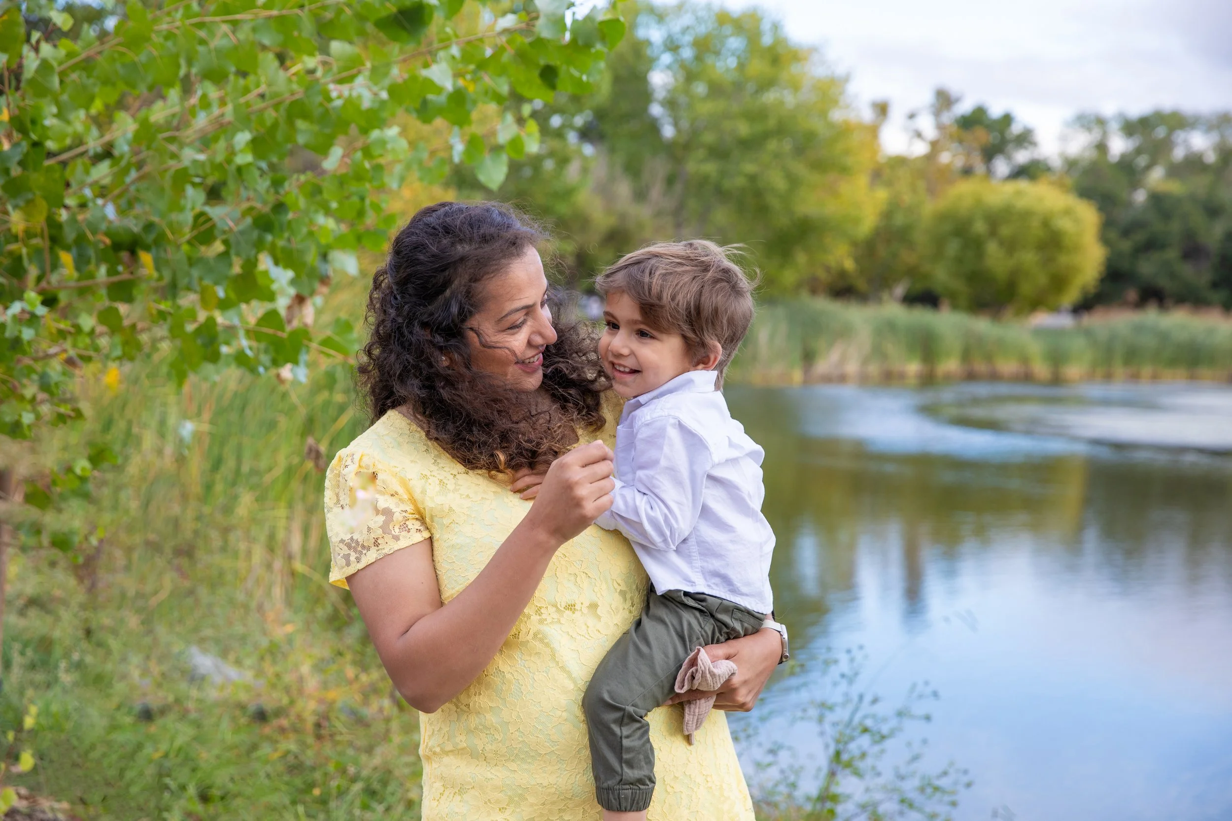 A woman and a young boy smiling at each other outdoors near a river, surrounded by trees and greenery.