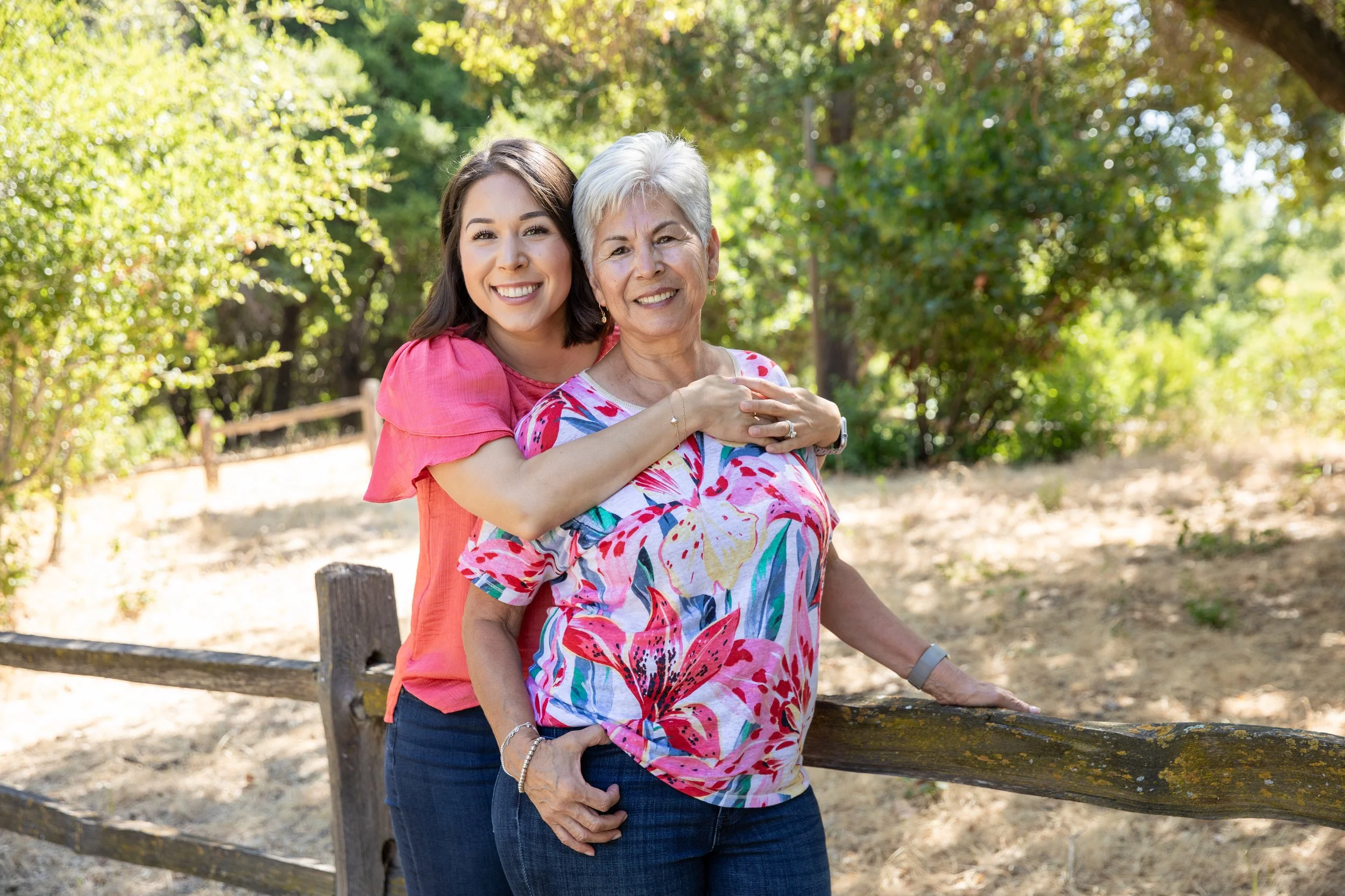 A young woman hugging an older woman from behind outdoors on a sunny day, with green trees and a wooden fence in the background.