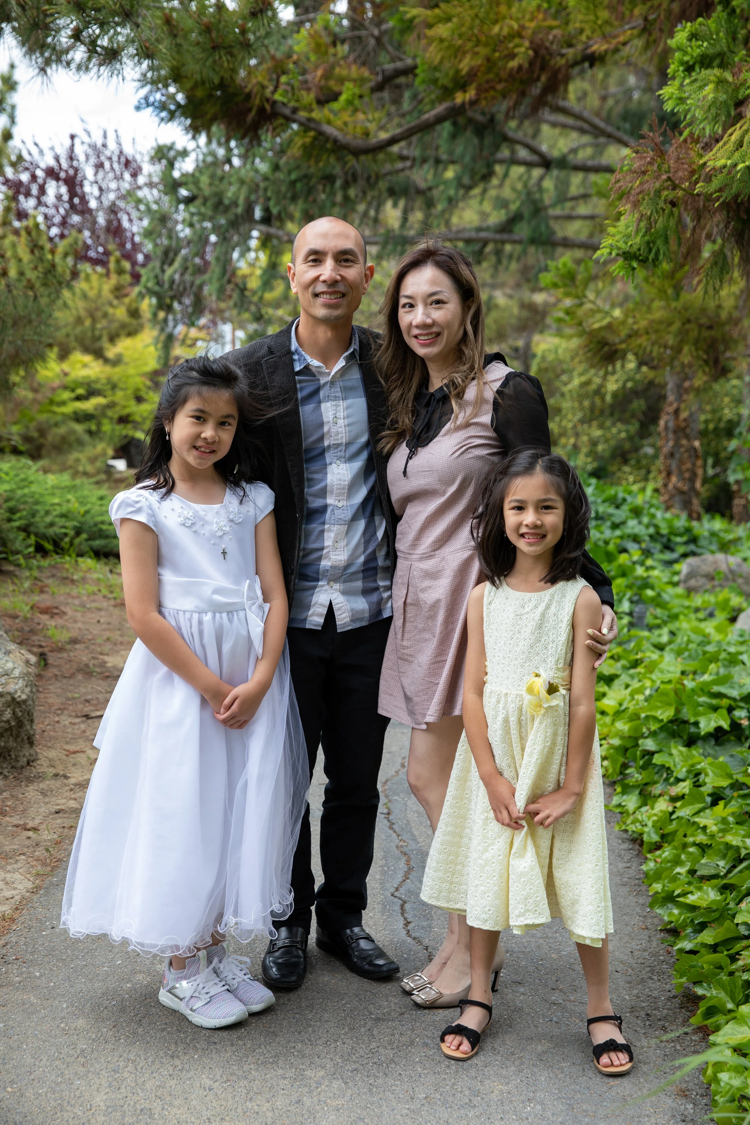 A family of four, including a man, woman, and two young girls, stands together outdoors on a paved path surrounded by green trees and foliage, smiling at the camera.