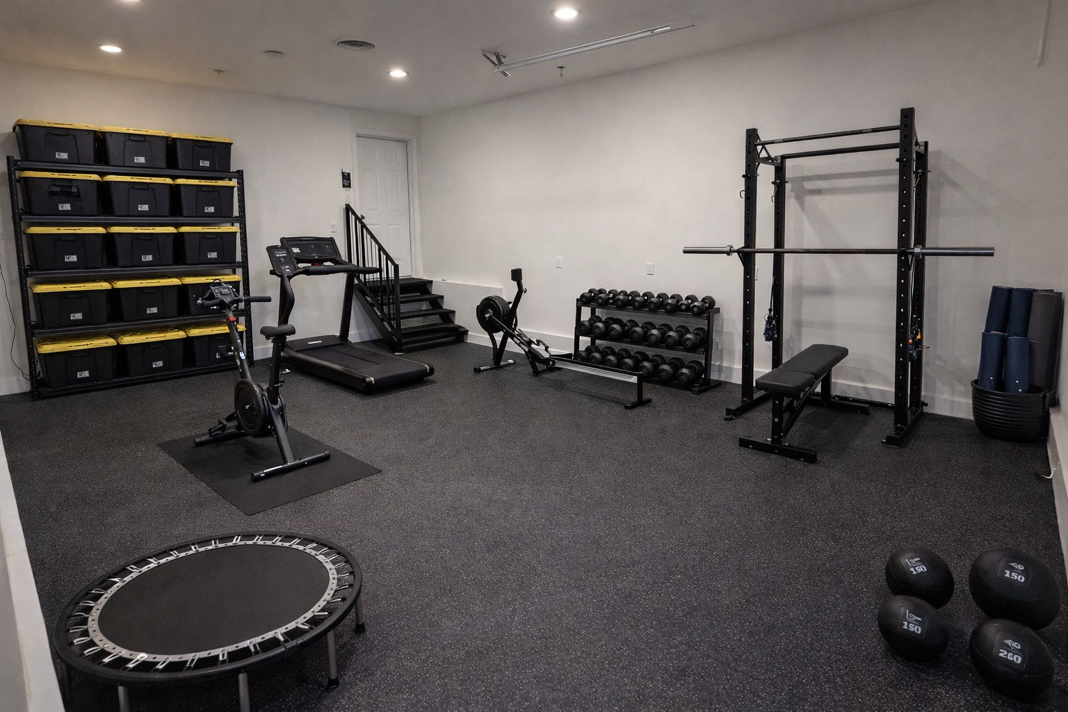 Home gym with small trampoline, exercise bike, treadmill, dumbbell rack with weights, weightlifting bench and barbell, resistance bands, foam rollers, and storage shelves for equipment, inside a well-lit room with black flooring.