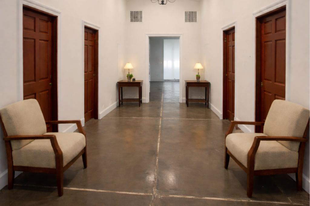Hallway with four wooden doors, two beige armchairs, and two small side tables with lamps, beige walls, concrete floor, and vents near the ceiling.
