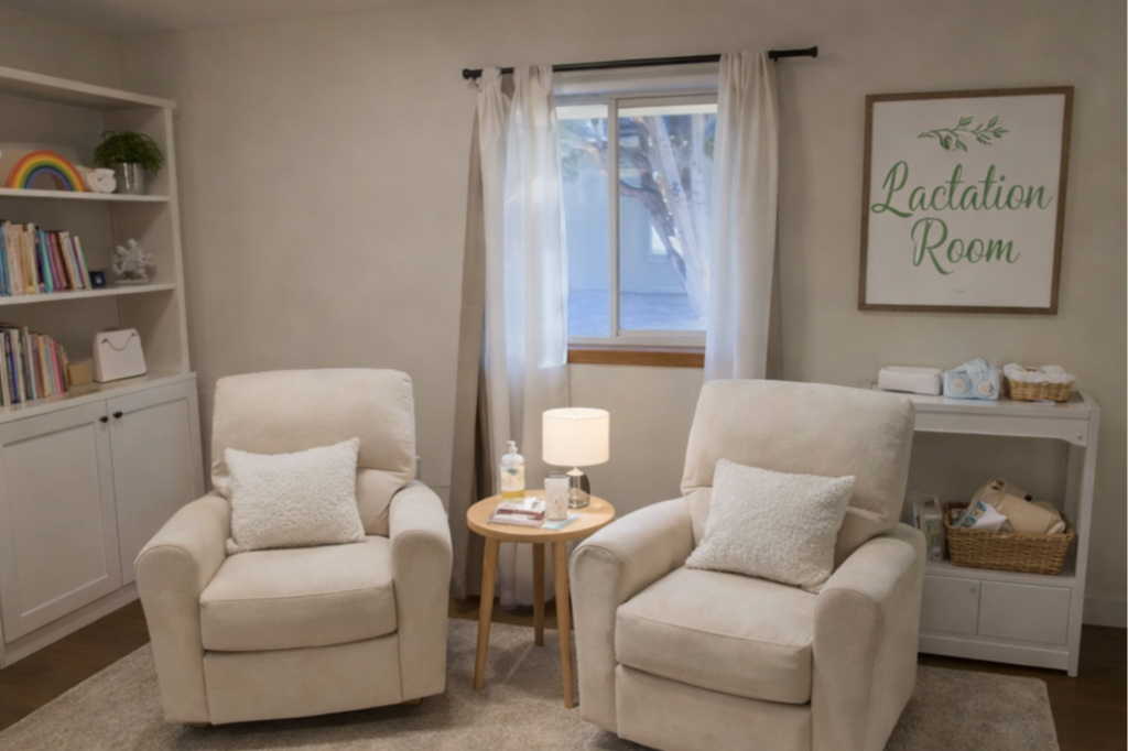 A cozy lactation room with two beige armchairs, a small wooden side table with a lamp, a window with white curtains, a white bookshelf filled with books and decorative items, and a sign that reads "Lactation Room" on the wall.