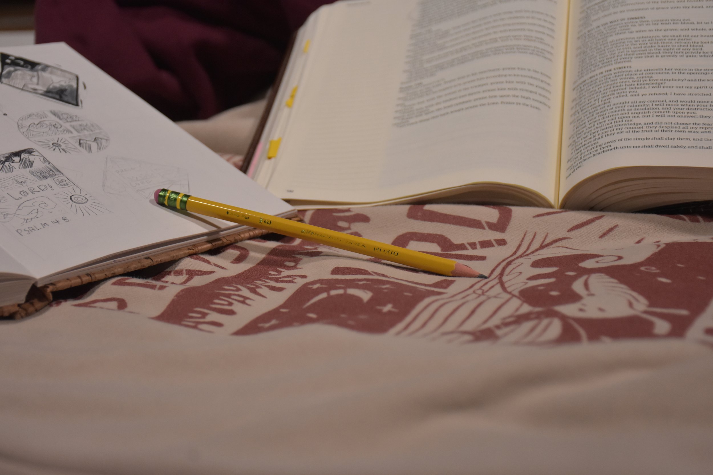 Open Bible, sketchbook with drawings, yellow pencil with pink eraser resting on a cloth with a graphic print.