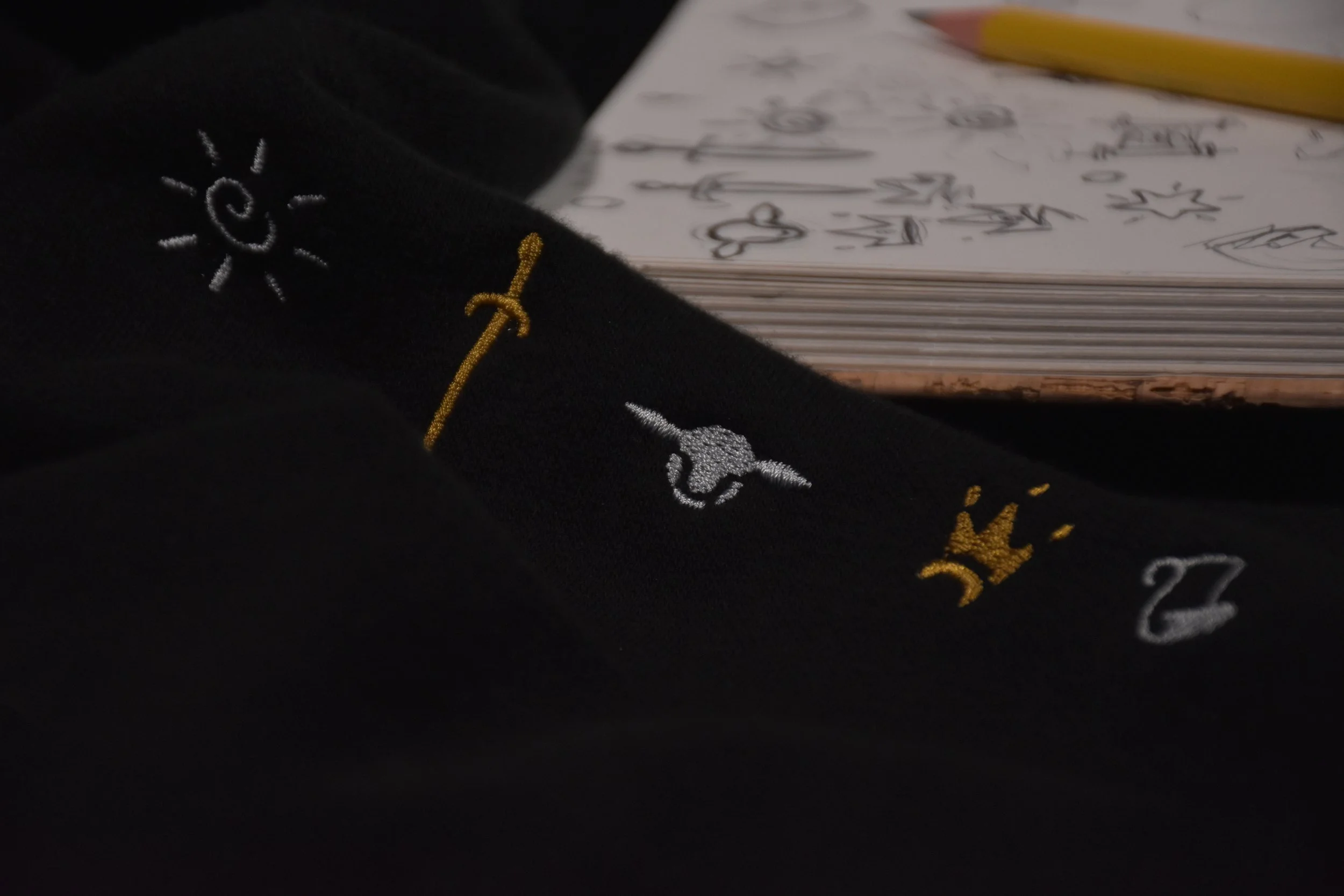 Close-up of a black fabric with embroidered symbols, including a sun, a lamb head, and a crown, near an open sketchpad with drawings.