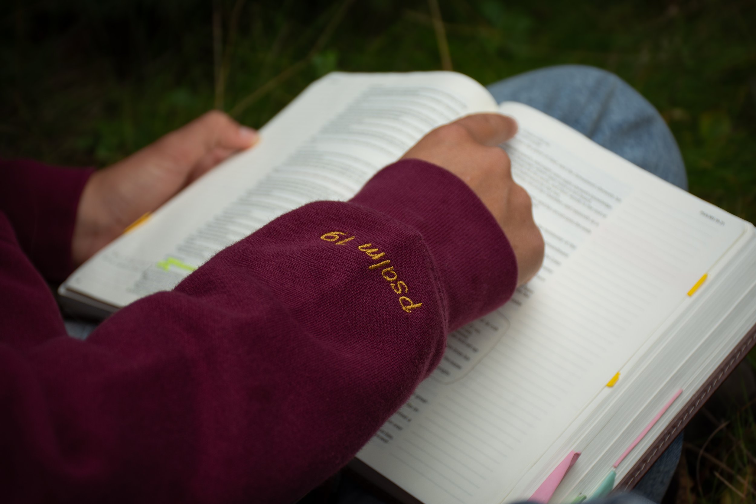 Person reading a Bible outdoors, wearing a maroon sweatshirt with yellow embroidered text reading Psalm 19 on the sleeve.