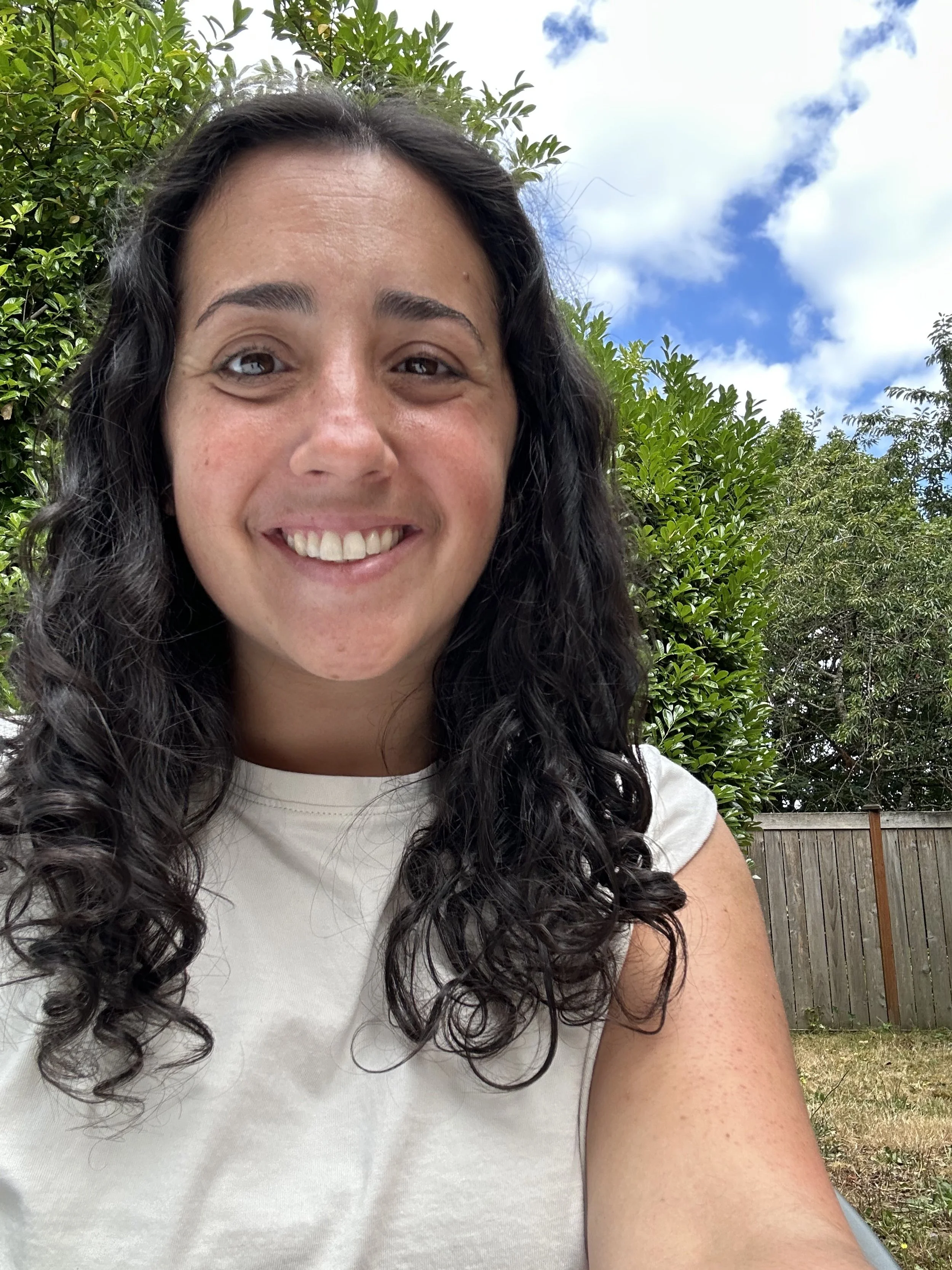 A woman with long curly dark hair taking a selfie outdoors, smiling, wearing a white shirt, with green trees, blue sky, and a wooden fence in the background.