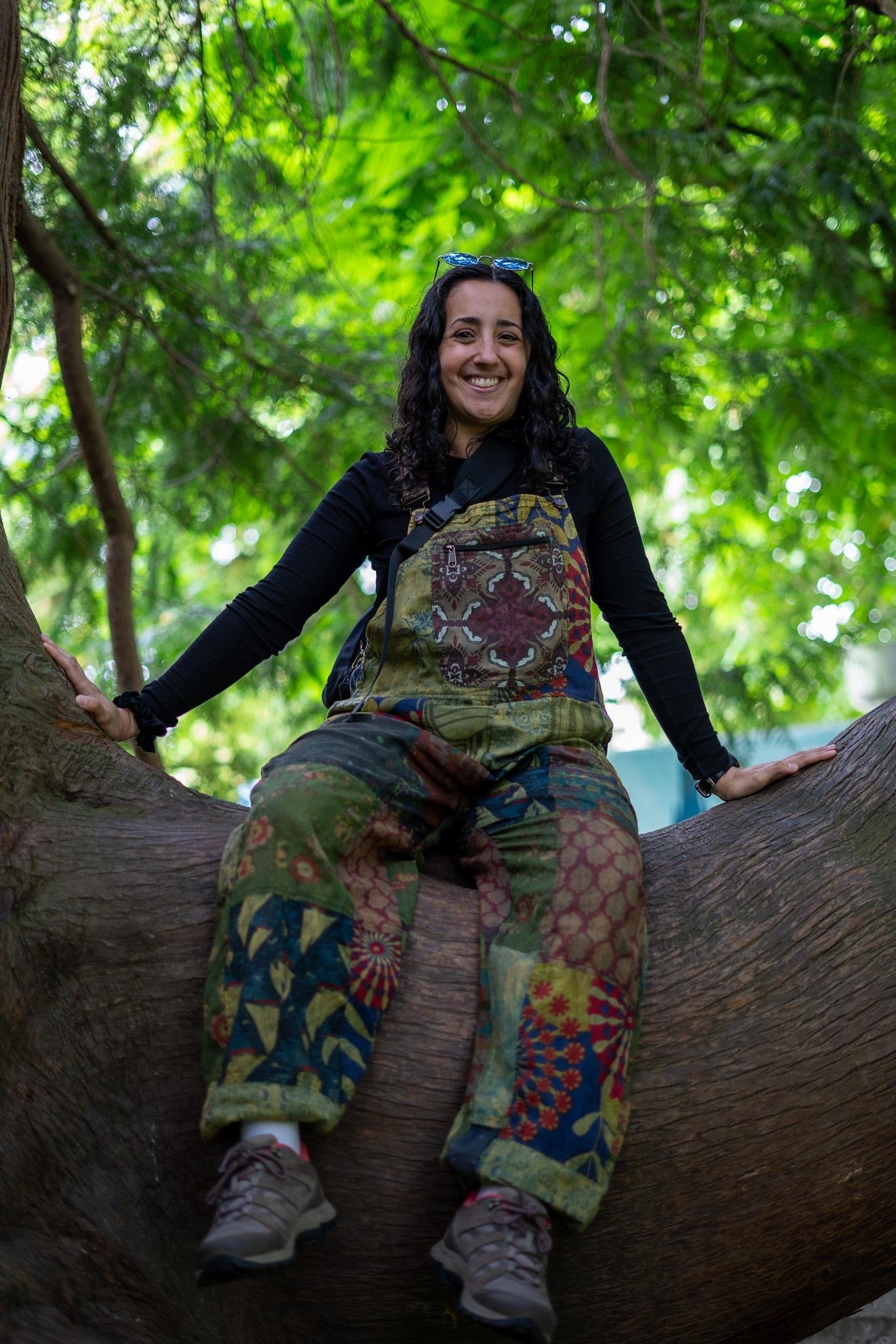 Woman sitting on a large tree branch in a forest, smiling and wearing glasses on her head, a backpack, patterned pants, and sneakers.