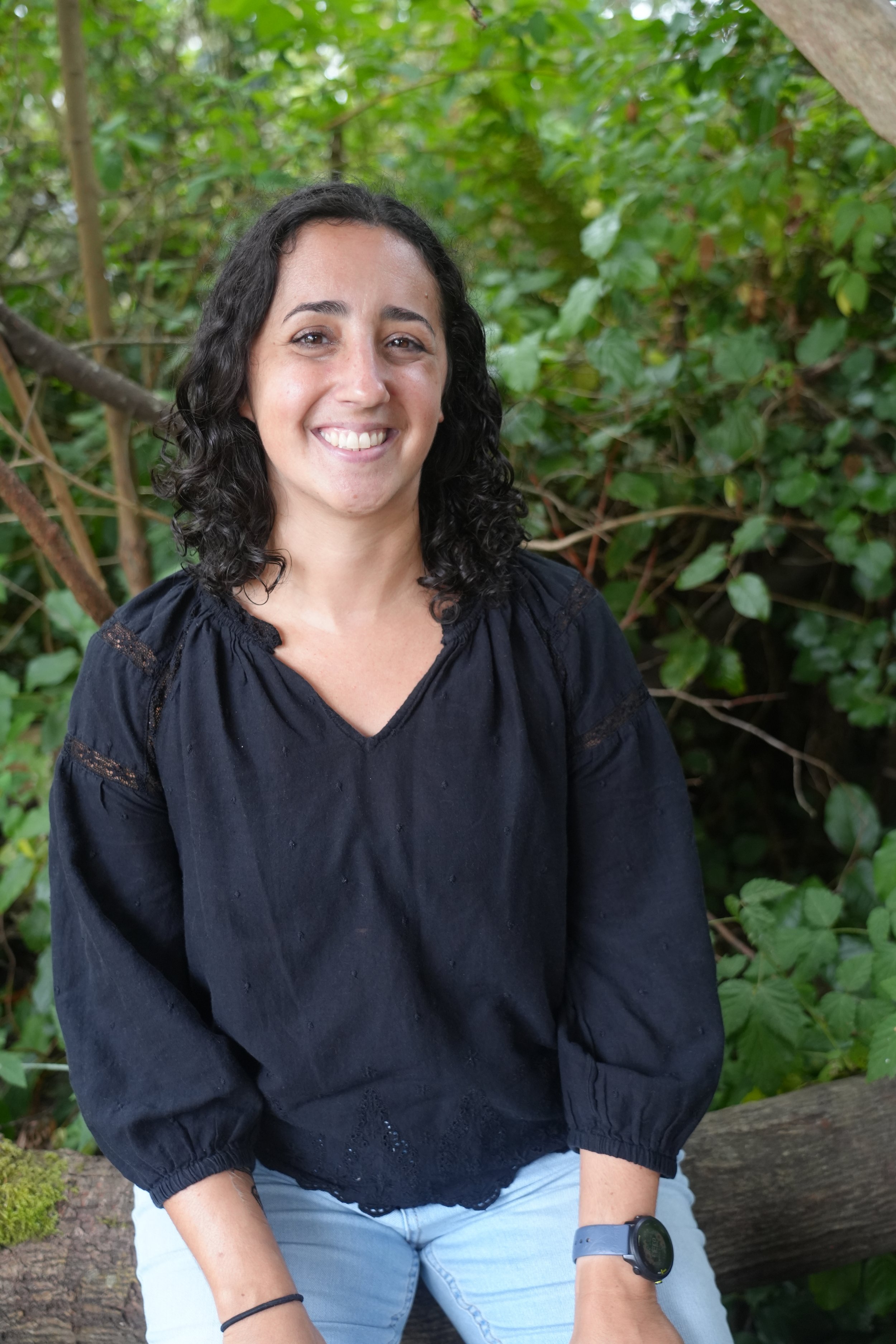 Smiling woman with shoulder-length dark curly hair wearing a black blouse and light blue jeans, seated on a fallen log surrounded by lush green foliage.