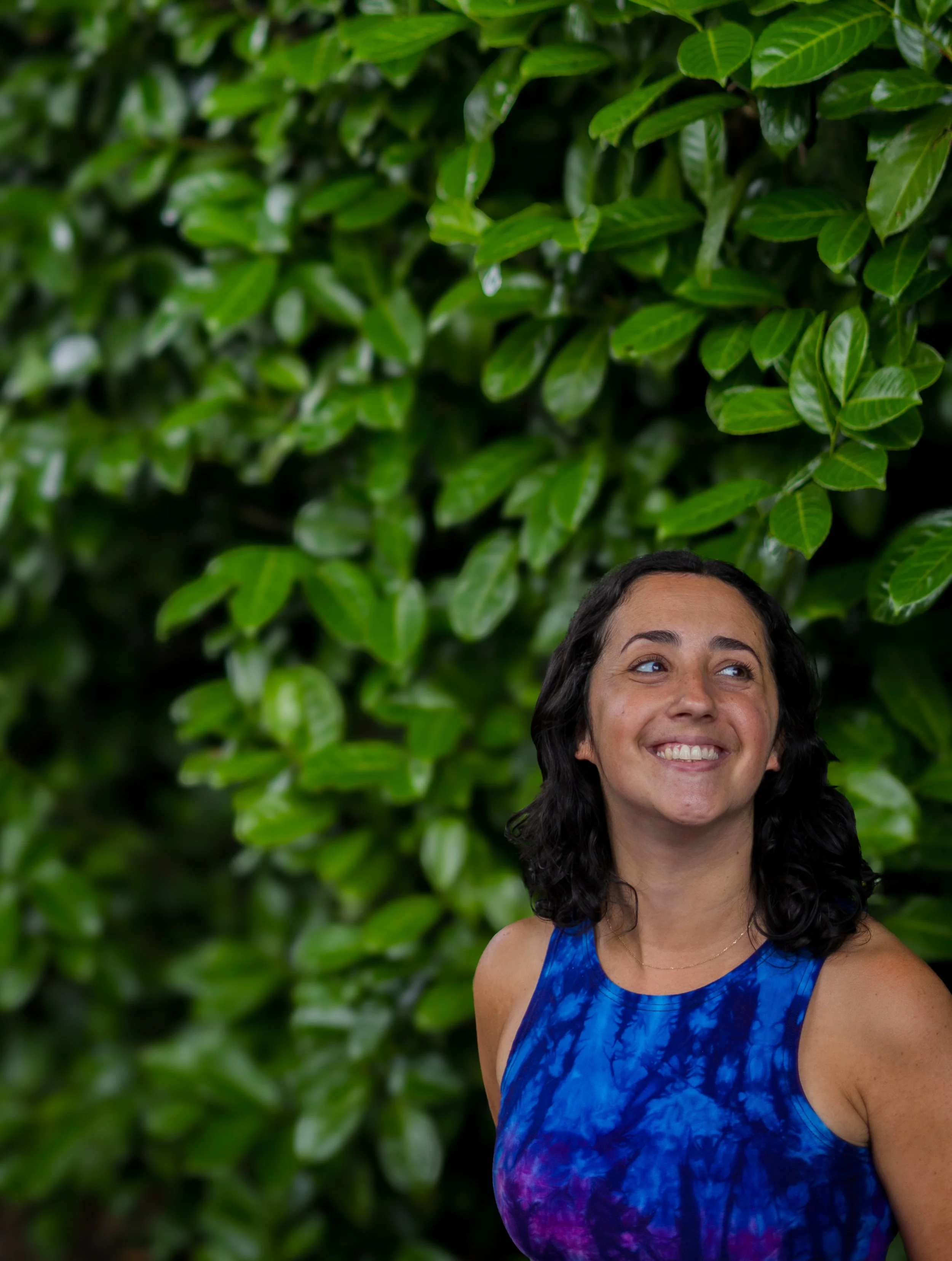 A woman with dark, wavy hair and a bright smile wearing a blue tie-dye sleeveless top, standing in front of a lush, green leafy background.