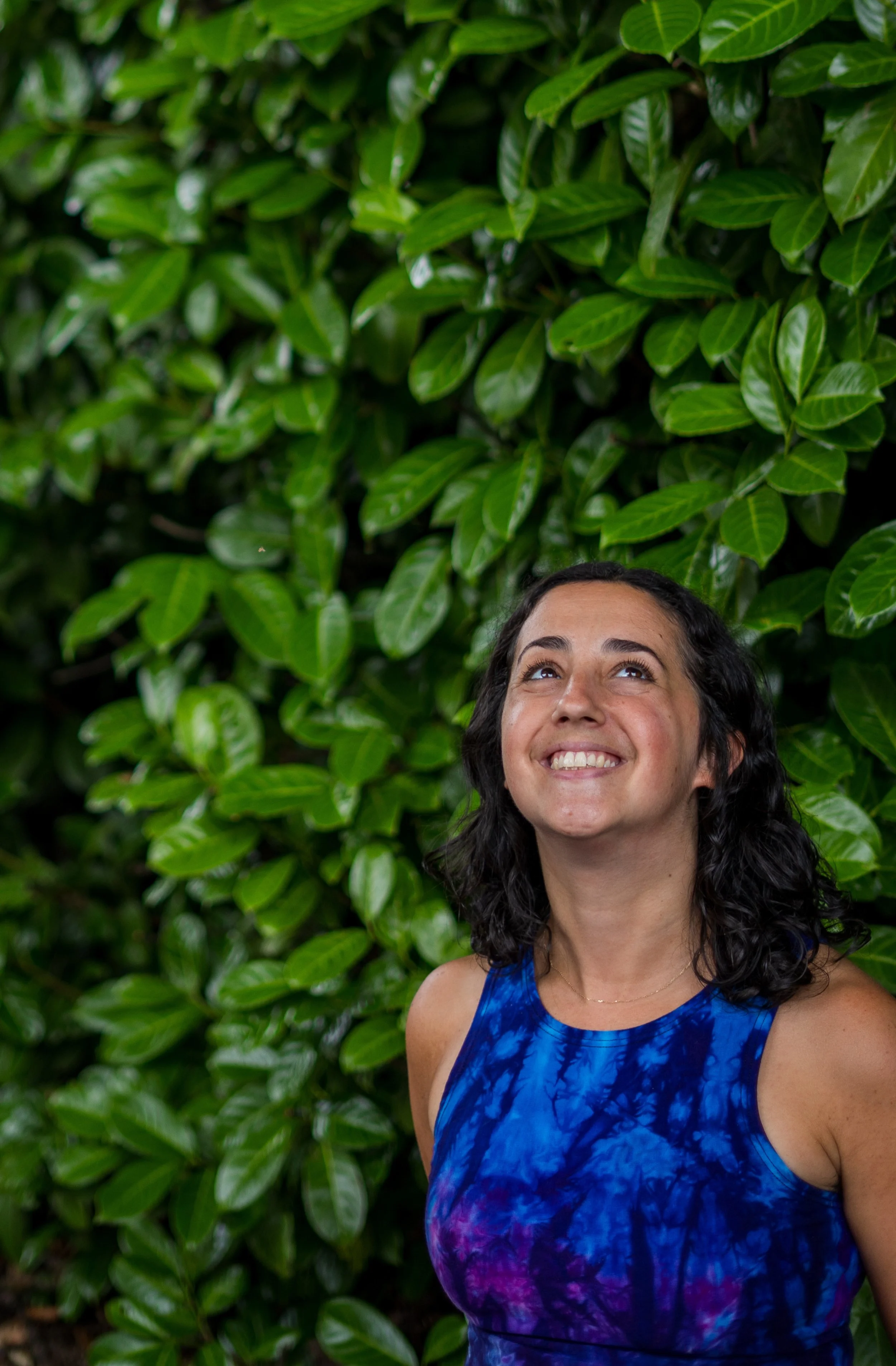 A woman with dark curly hair smiling and looking upward, wearing a sleeveless blue tie-dye dress, standing in front of a lush green leafy background.