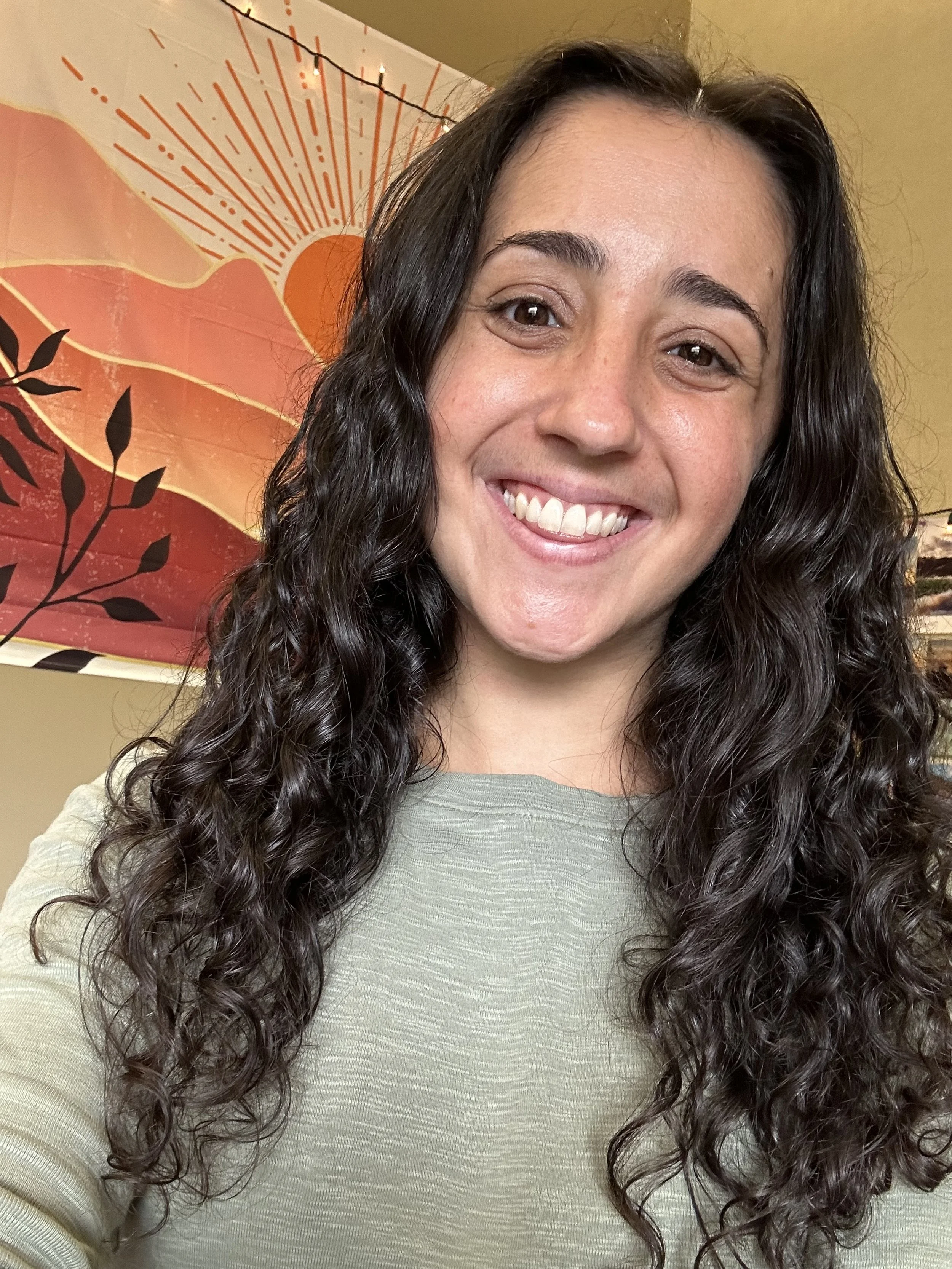 A woman with long, curly dark hair smiling at the camera, standing in front of a colorful wall art with a sunset scene and black branch silhouette.