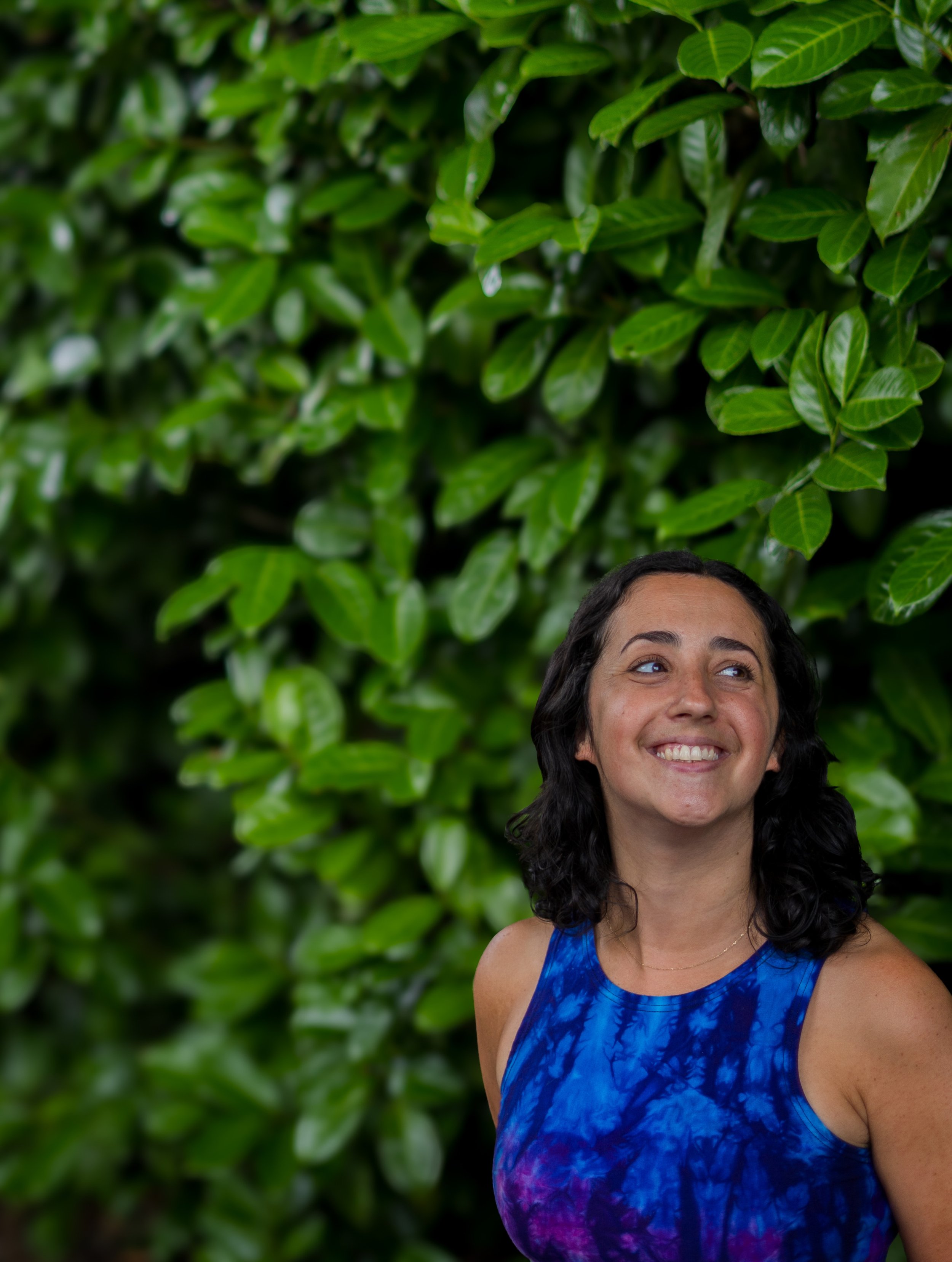 The image features a smiling woman with dark hair and light skin, wearing a sleeveless blue patterned top. She is set against a lush green leafy background with vibrant foliage.