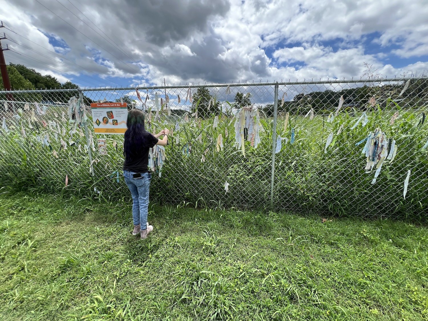 Visitor tying fabric to the fence as summer vegetation grows taller around the site.
