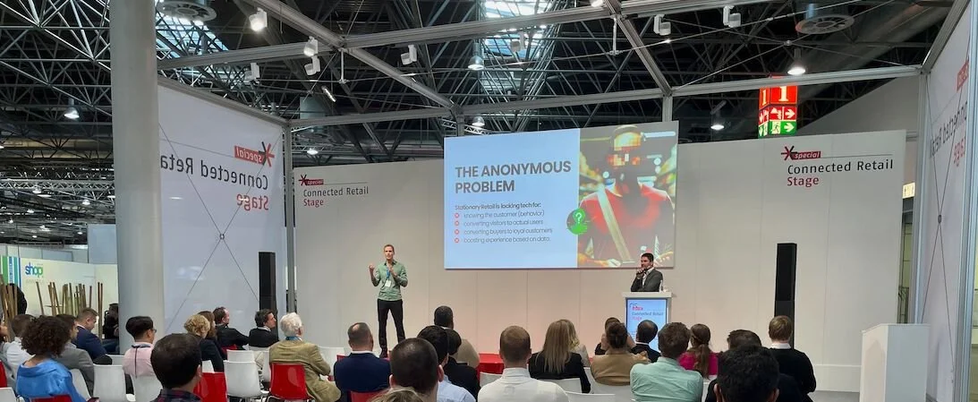 Eine Konferenzhalle mit einem Publikum, das zwei Sprecher auf der Bühne sieht, die über das Thema 'Das anonyme Problem' sprechen. Die Bühne hat einen Bildschirm mit einer Präsentation und der Text 'Connected Retail Stage'.