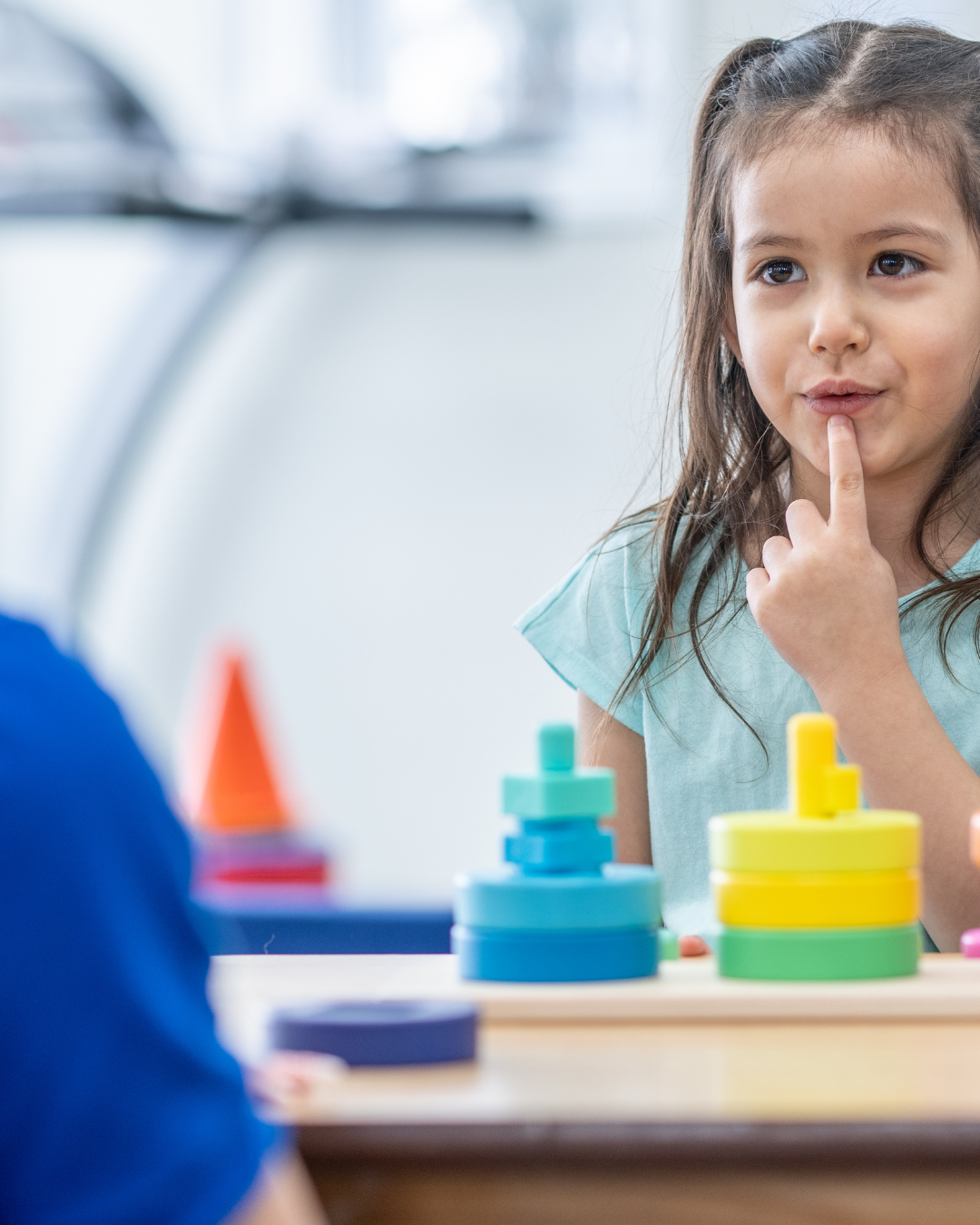 A young girl in a teal shirt thinks deeply, touching her chin, while playing with colorful stacking rings at a table in a bright, playful space.