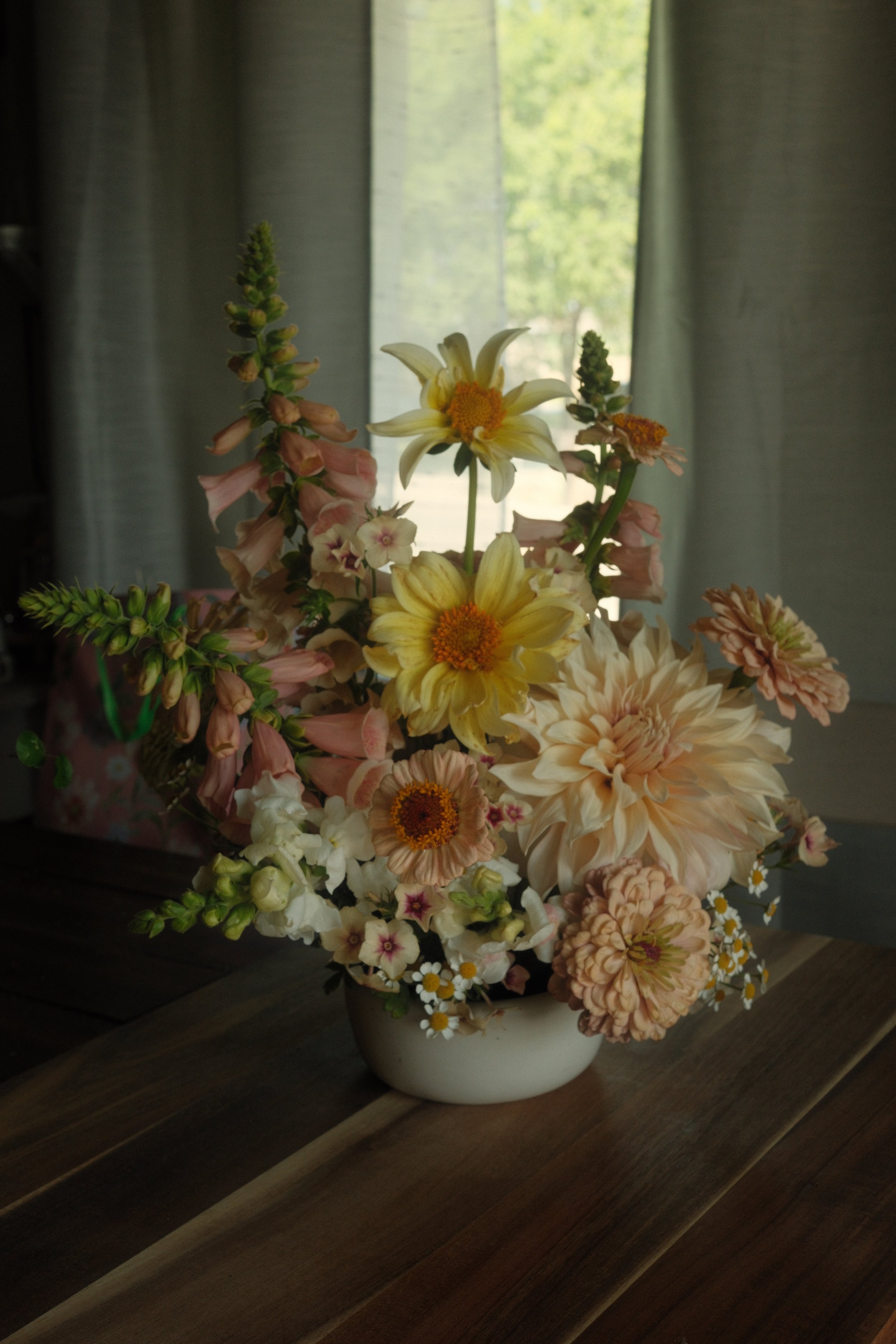 A bouquet of various flowers in a white vase on a wooden table, with curtains and a window showing green trees outside in the background.