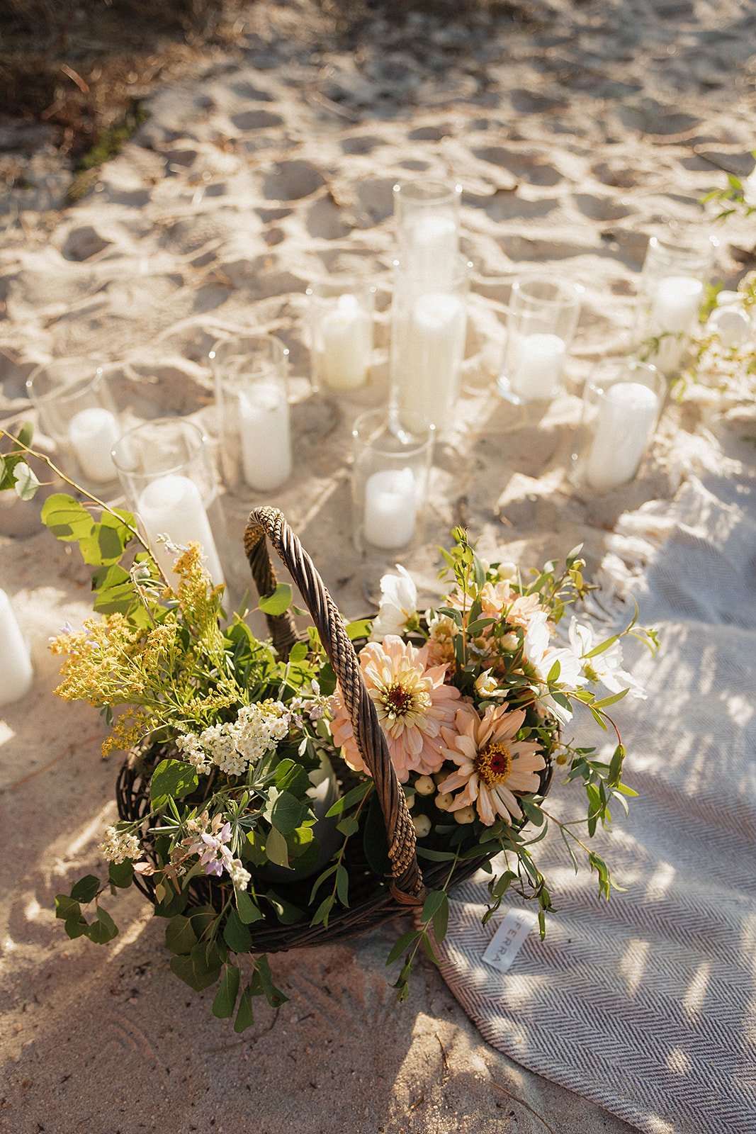 A wicker basket filled with light pink and white flowers, decorated with green leaves, placed on sandy ground next to a fabric cloth, with several white candles in glass holders in the background, outdoors.