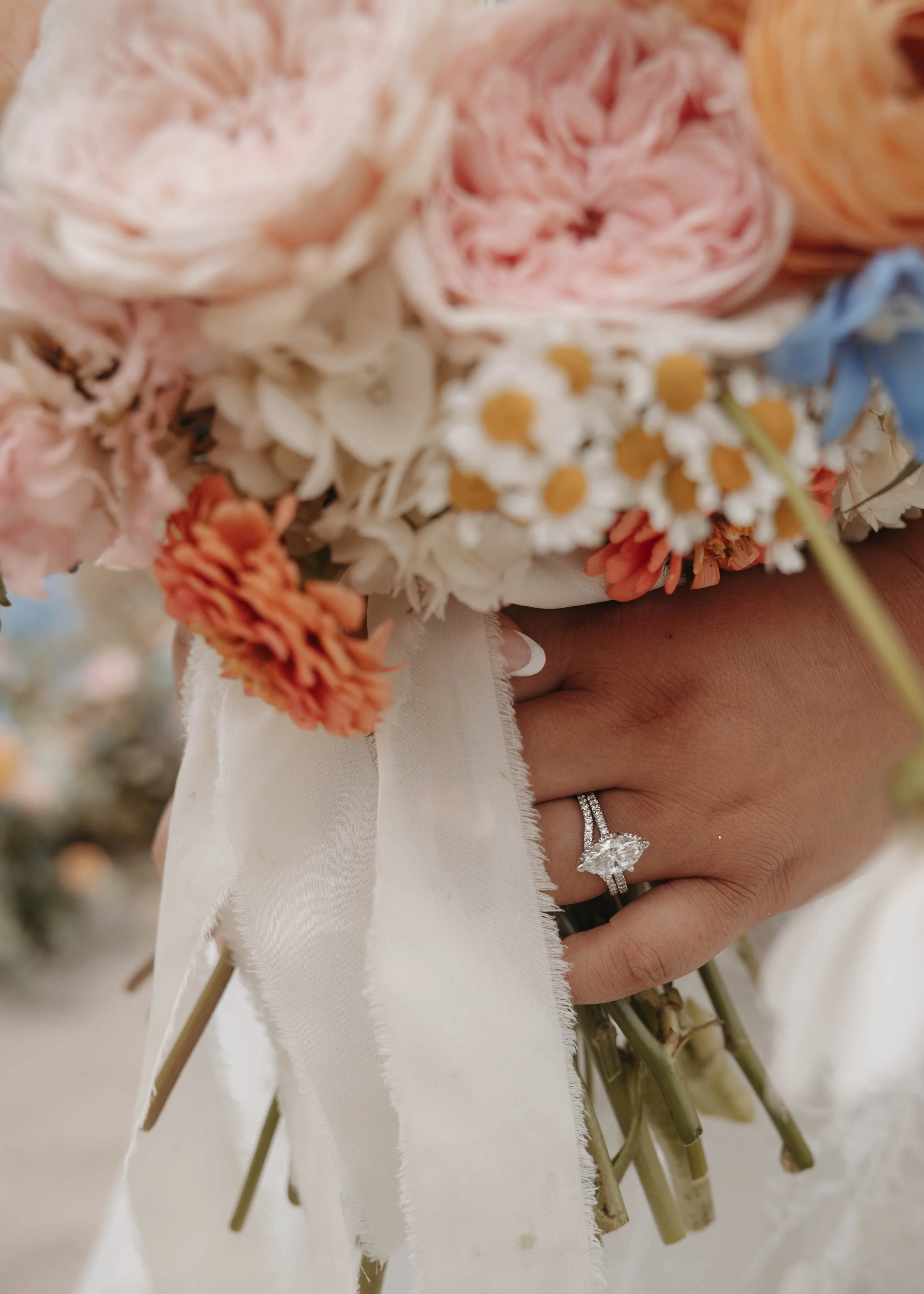 Close-up of a hand holding a wedding bouquet of pink, white, orange, yellow, and blue flowers, with a diamond engagement ring on the ring finger.