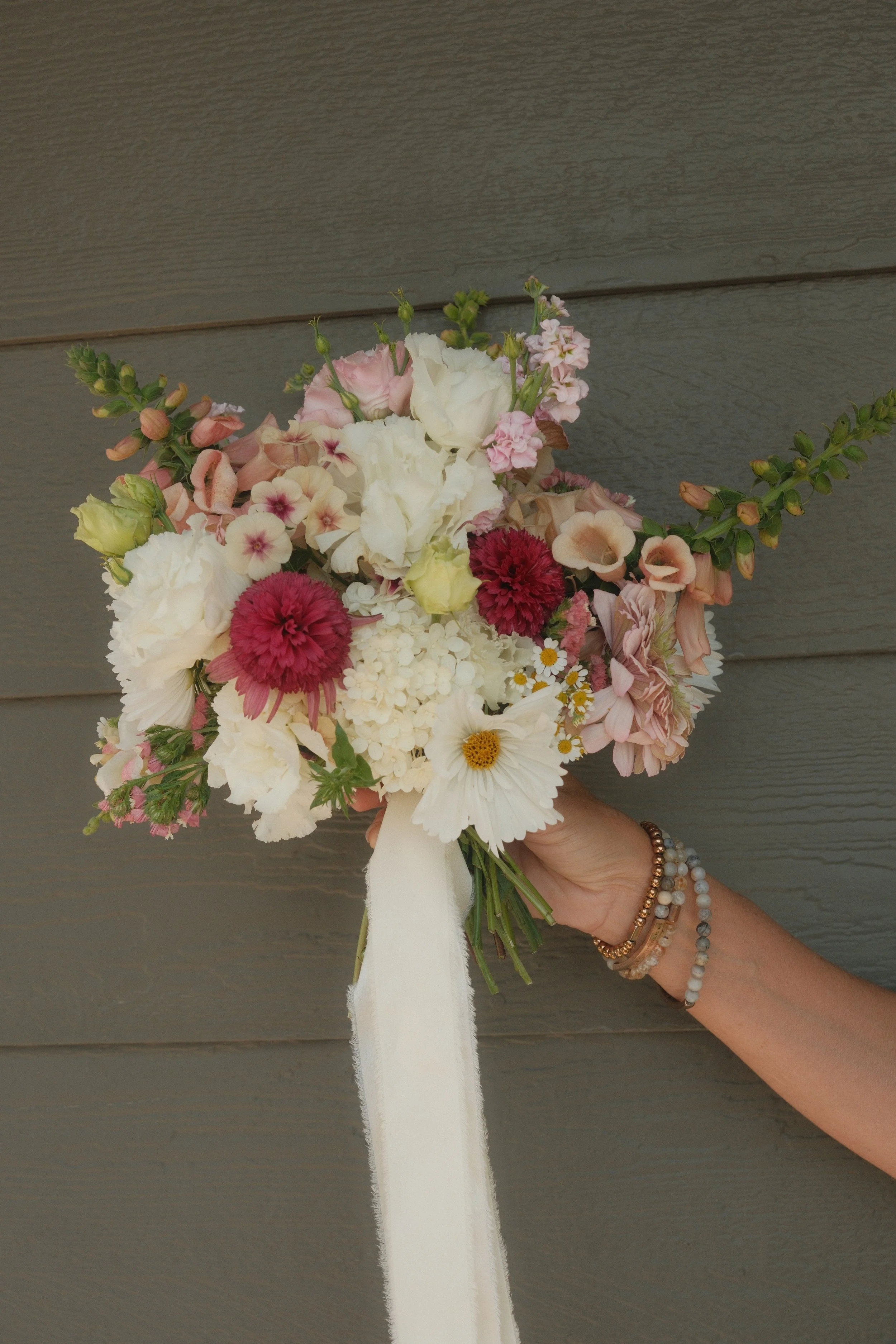 A hand holding a bouquet of pink, white, and burgundy flowers with a white ribbon, set against a gray wooden wall.