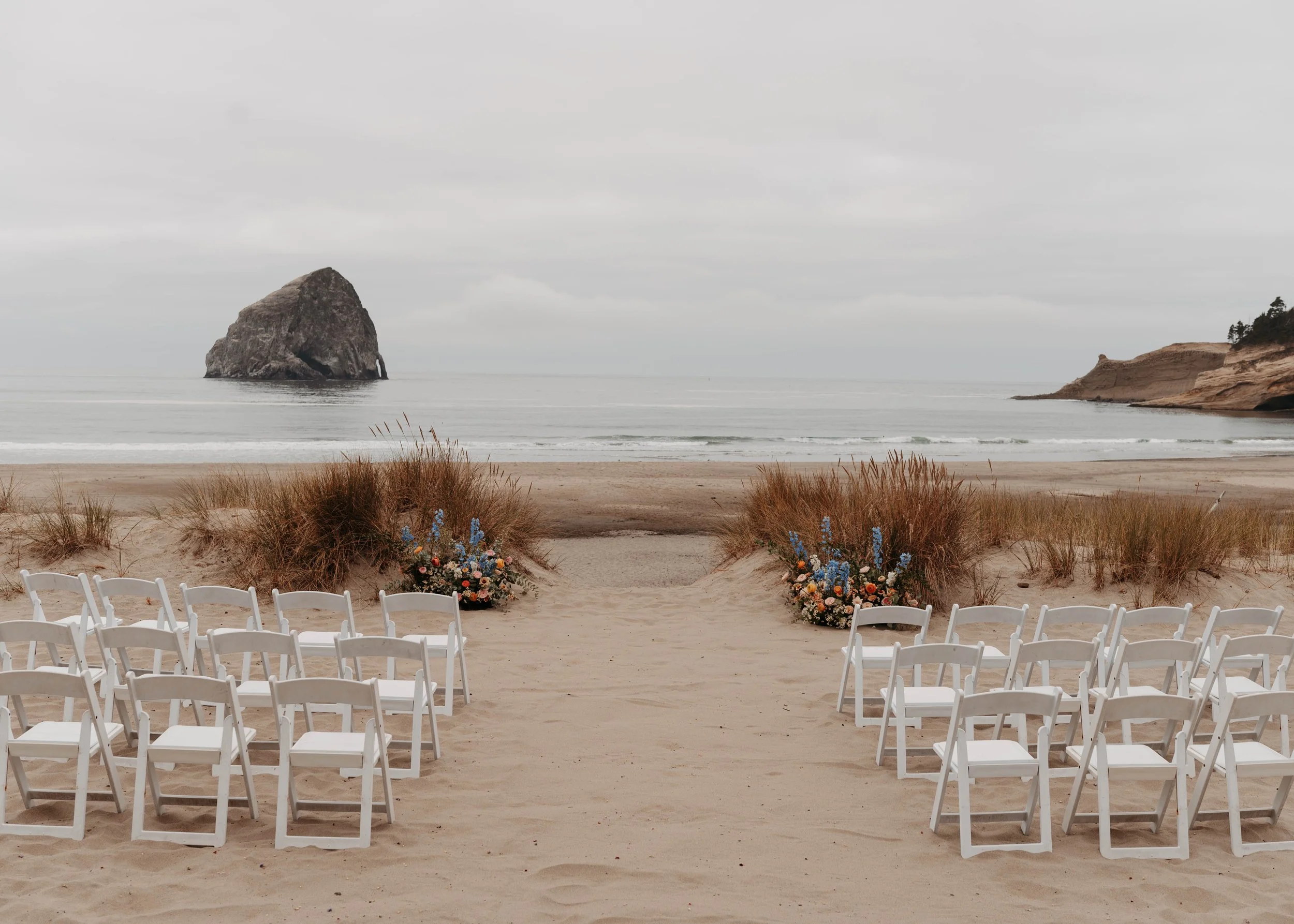 Empty white chairs arranged in rows on a sandy beach facing the ocean with rocks and cliffs in the background, decorated with flower arrangements and tall grasses.