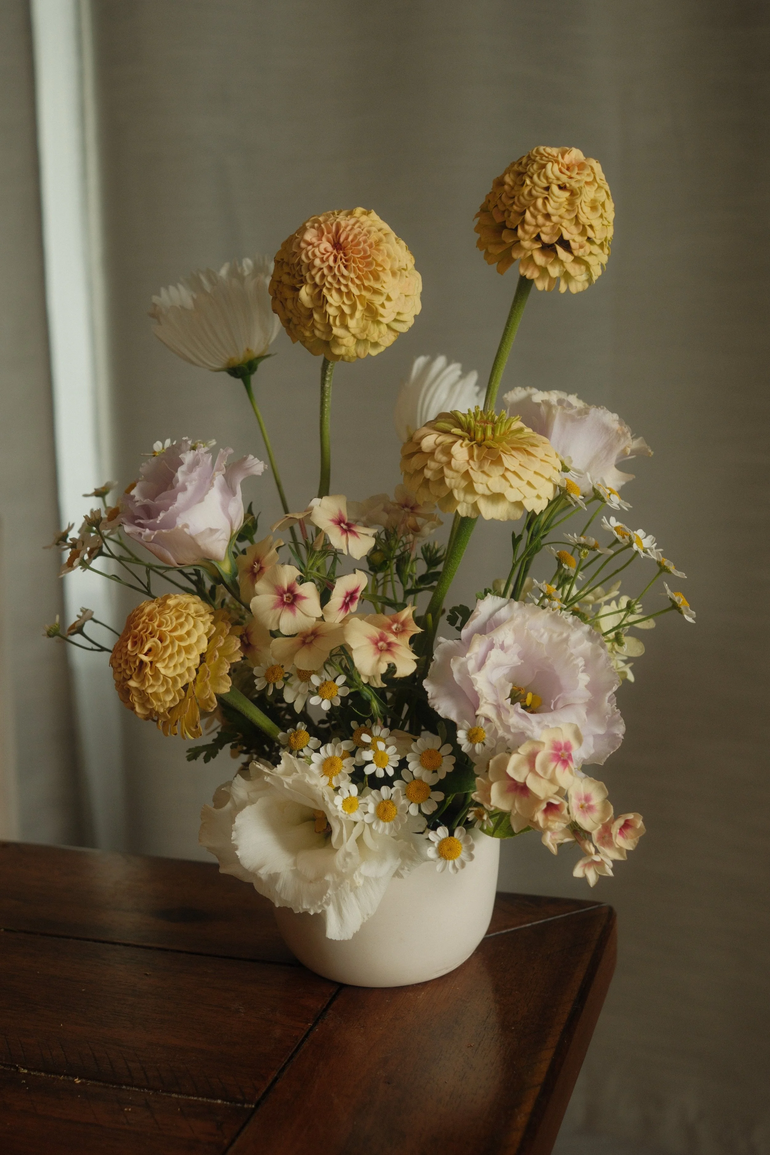 A white vase with a colorful variety of flowers including yellow, pink, white, and purple blooms, placed on a wooden surface.