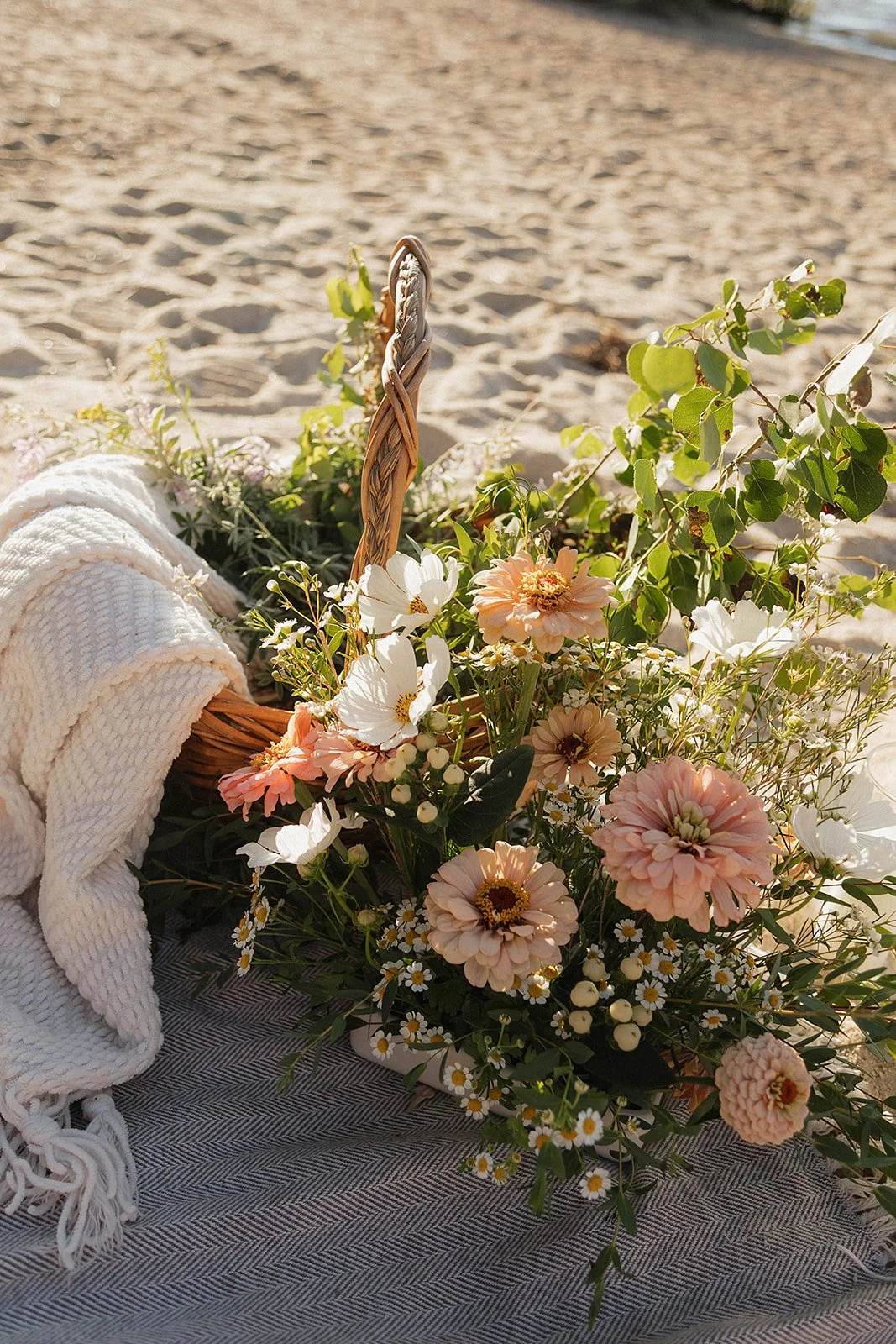 A basket filled with pink, white, and cream flowers, resting on a gray fabric with a rolled white towel nearby, on a sandy beach.