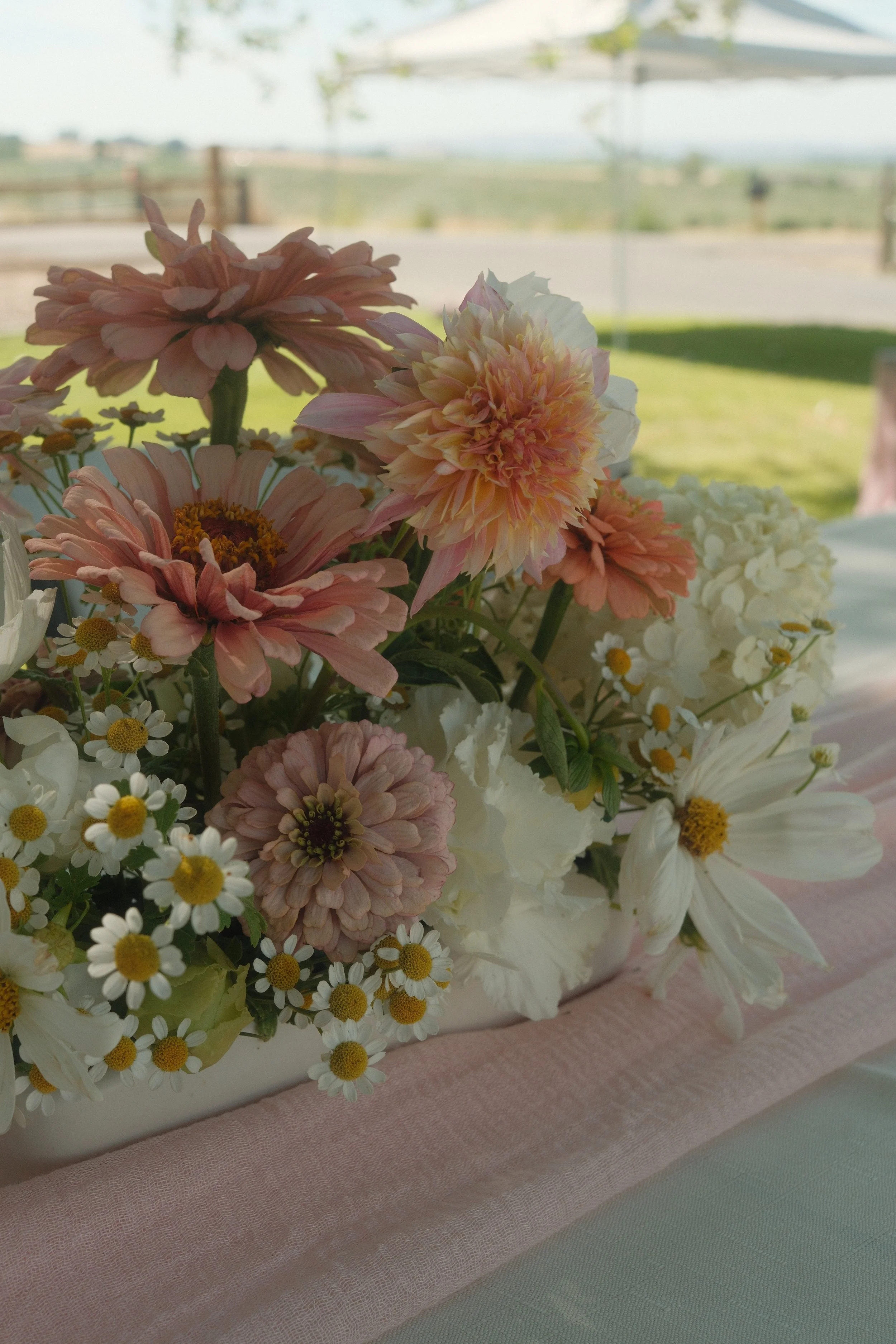A floral centerpiece with pink, peach, white, and yellow flowers on a pink table runner, outdoors with a deck and landscape in the background.
