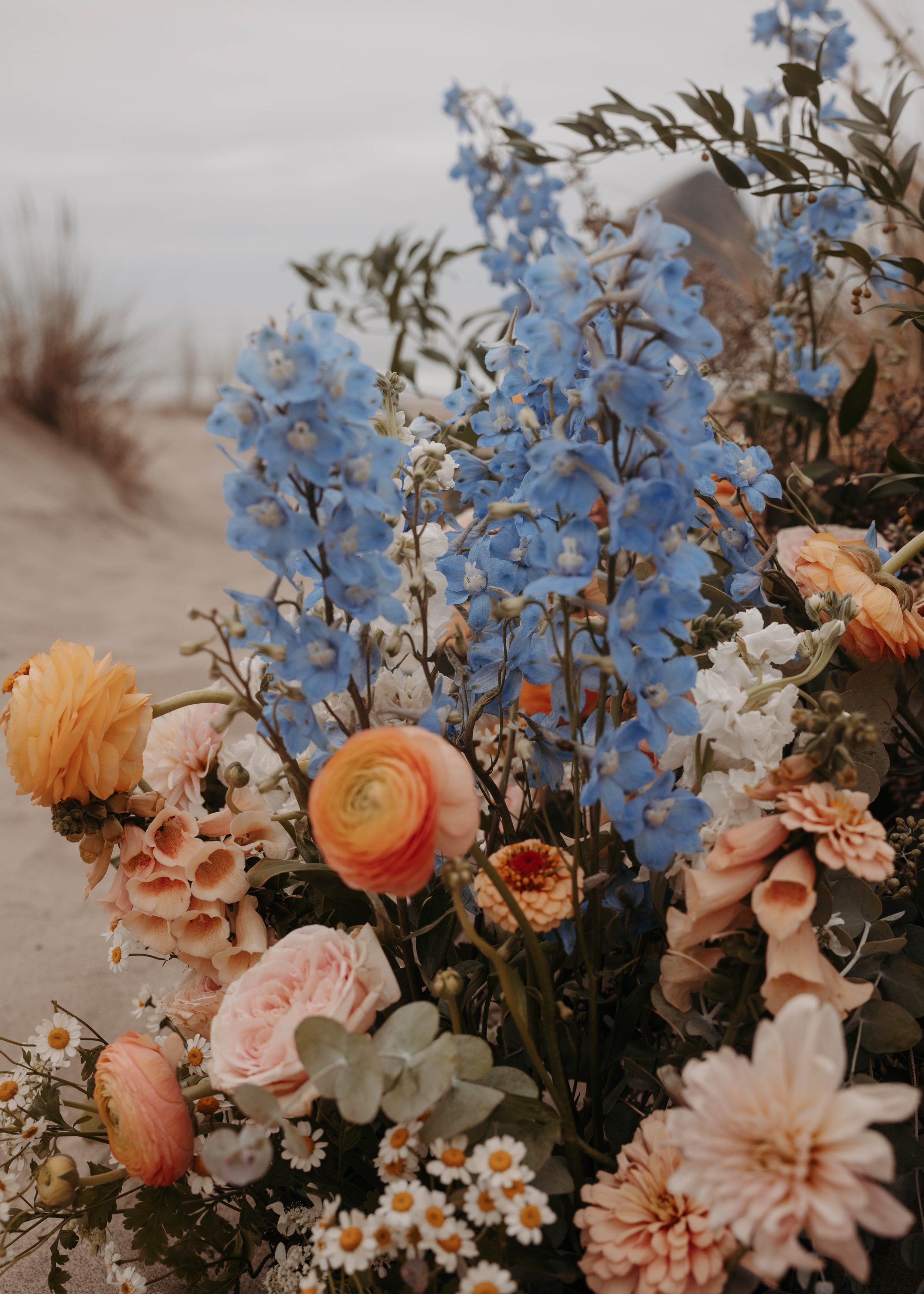 A bouquet of various flowers including blue delphiniums, pink ranunculus, white daisies, and other mixed blossoms on a sandy beach with dune grass in the background.