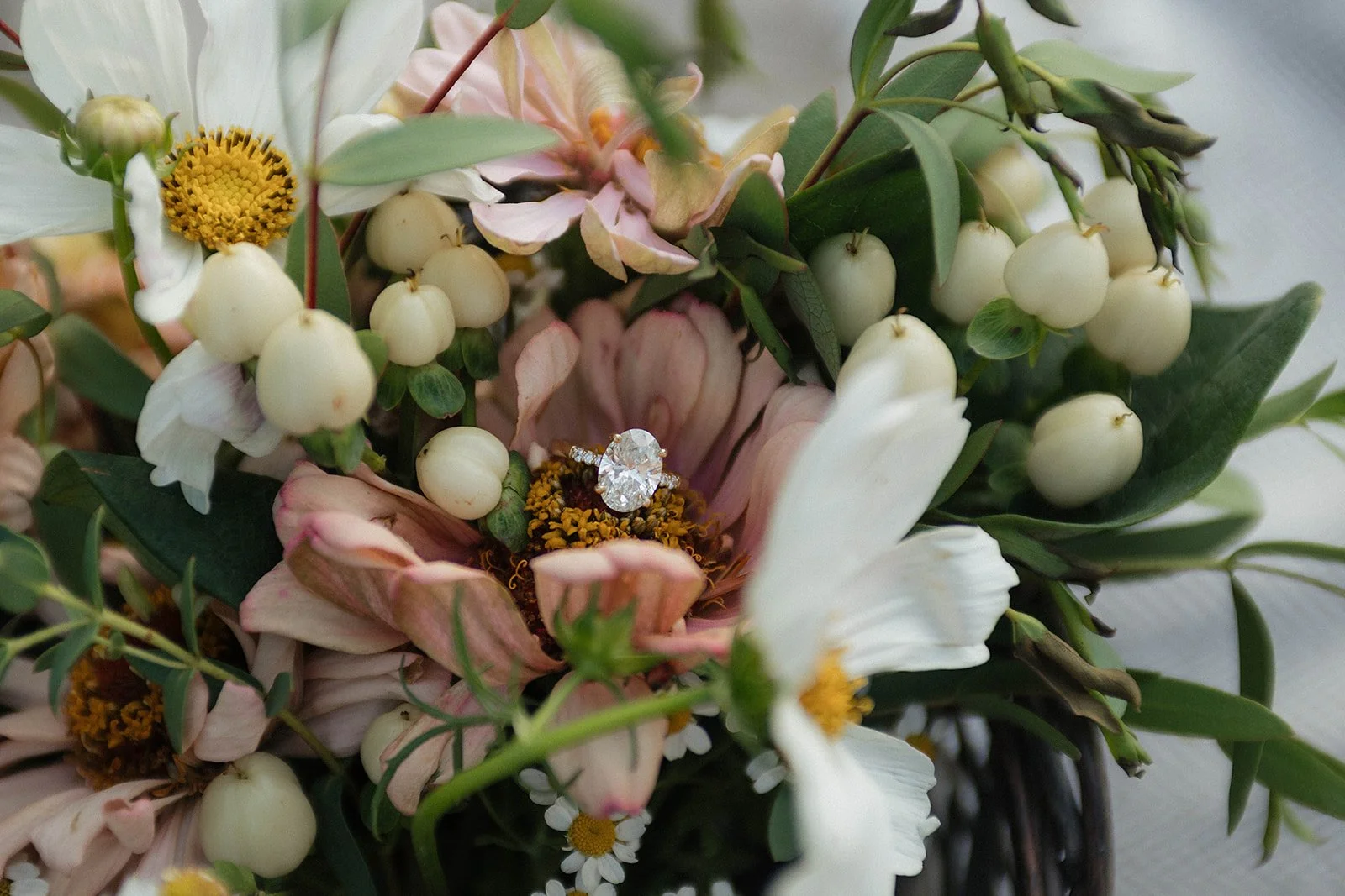 A close-up of a floral bouquet with white and pink flowers, green leaves, and berries, with a diamond ring placed on one of the flowers.