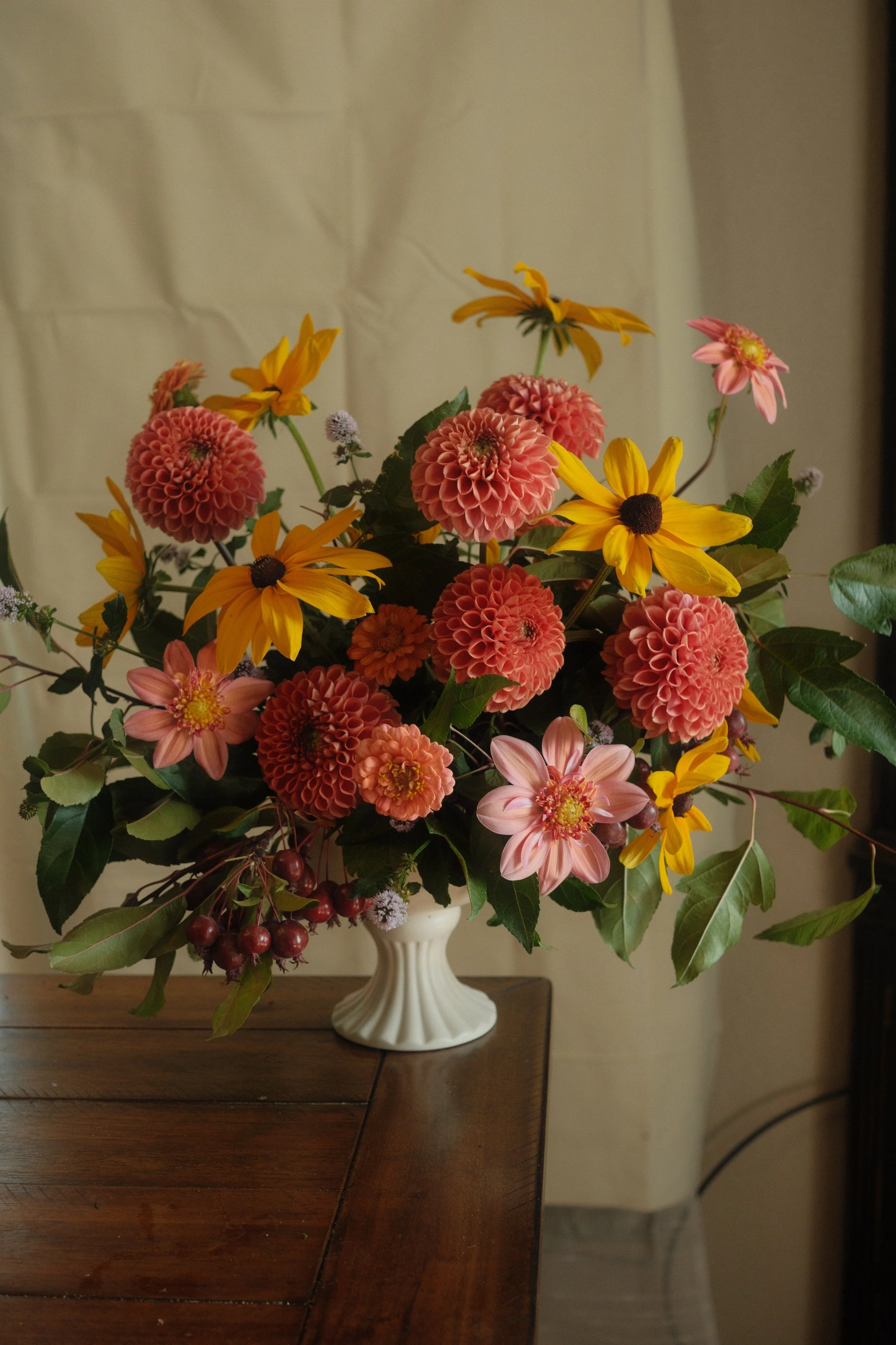 A bouquet of mixed flowers including pink dahlias, yellow daisies, and pink zinnias in a white vase on a wooden table.