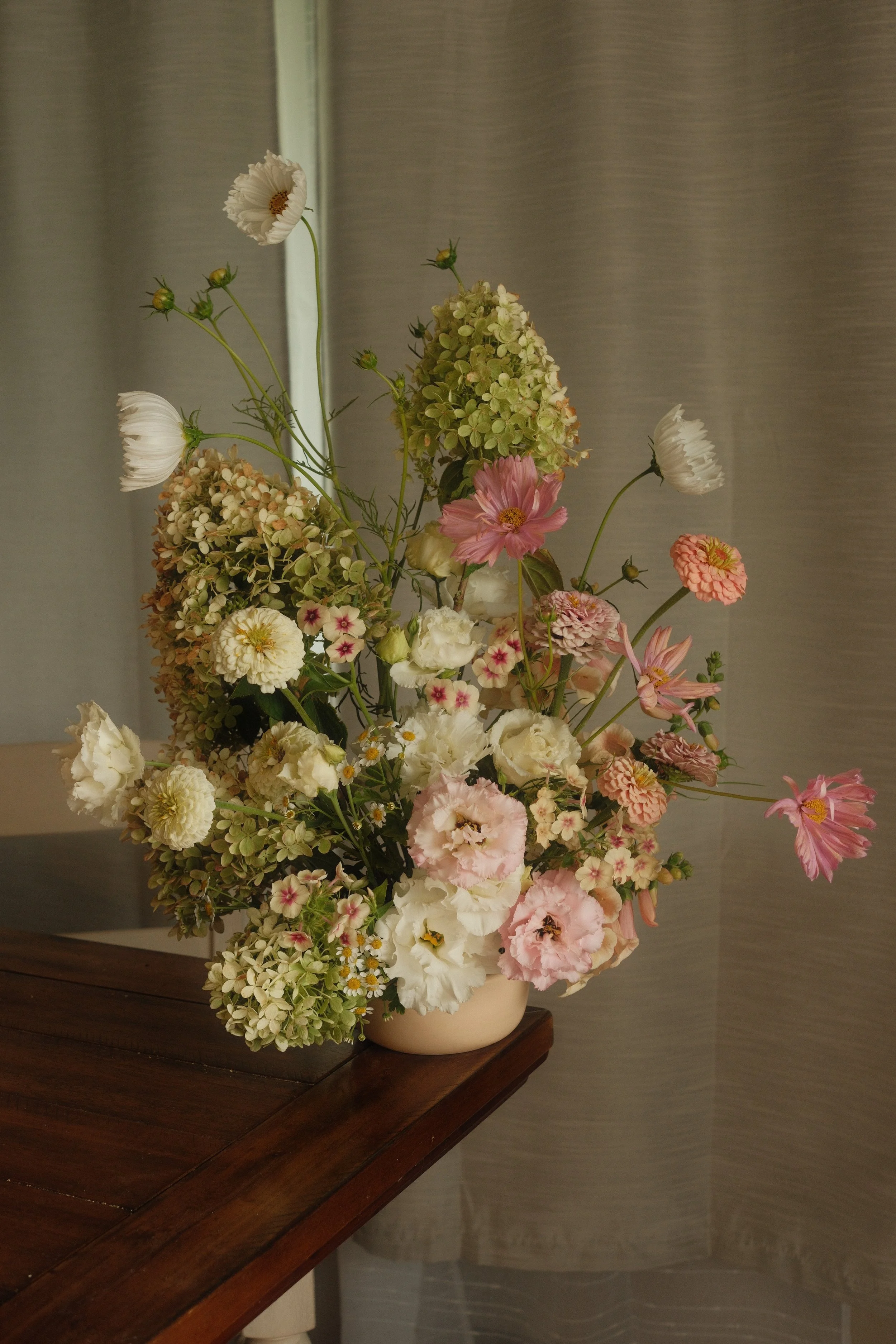 A bouquet of mixed flowers in a beige vase on a wooden table, with white, pink, and green blossoms.