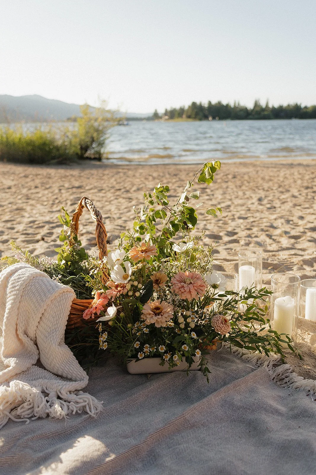A picnic setup on a sandy beach with a floral arrangement, white candles in glass holders, a white blanket, and a woven basket, overlooking a body of water with trees and mountains in the background.