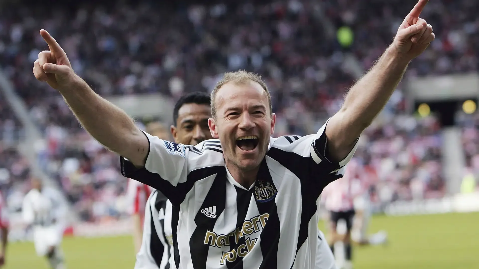A joyful soccer player wearing a black and white striped jersey with 'northern rocks' logo celebrates on the field with arms raised, smiling, with a stadium full of spectators in the background.