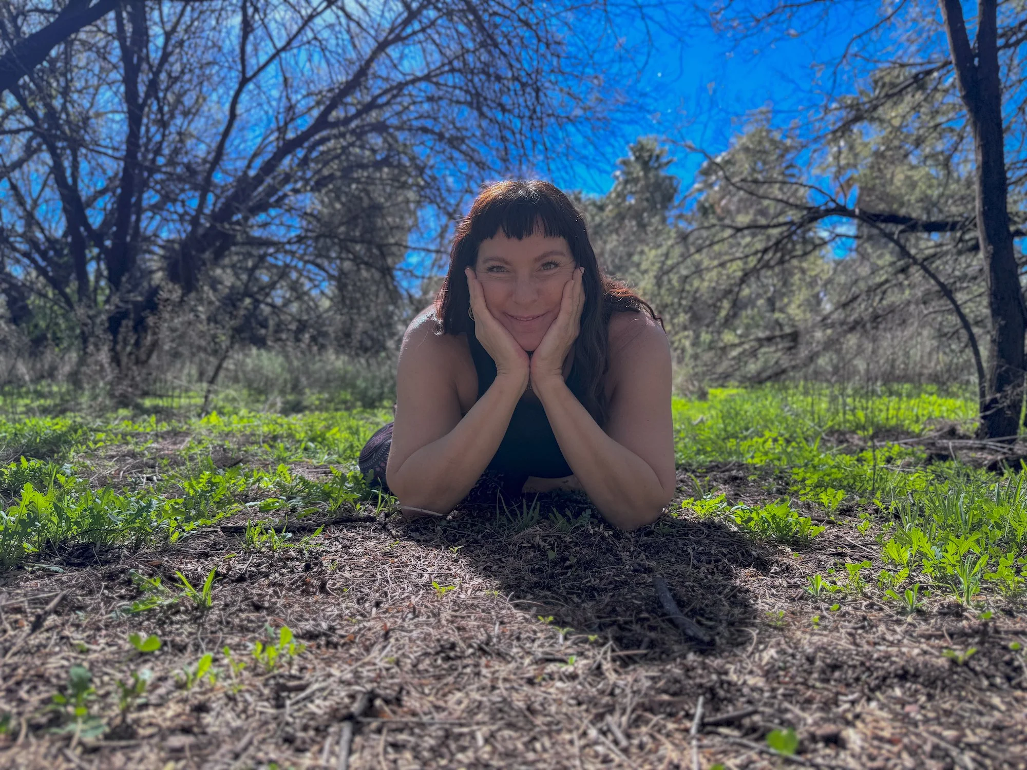 A woman with dark hair and bangs lying on her stomach on the ground, resting her face in her hands, smiling, surrounded by green plants and trees under a bright blue sky.