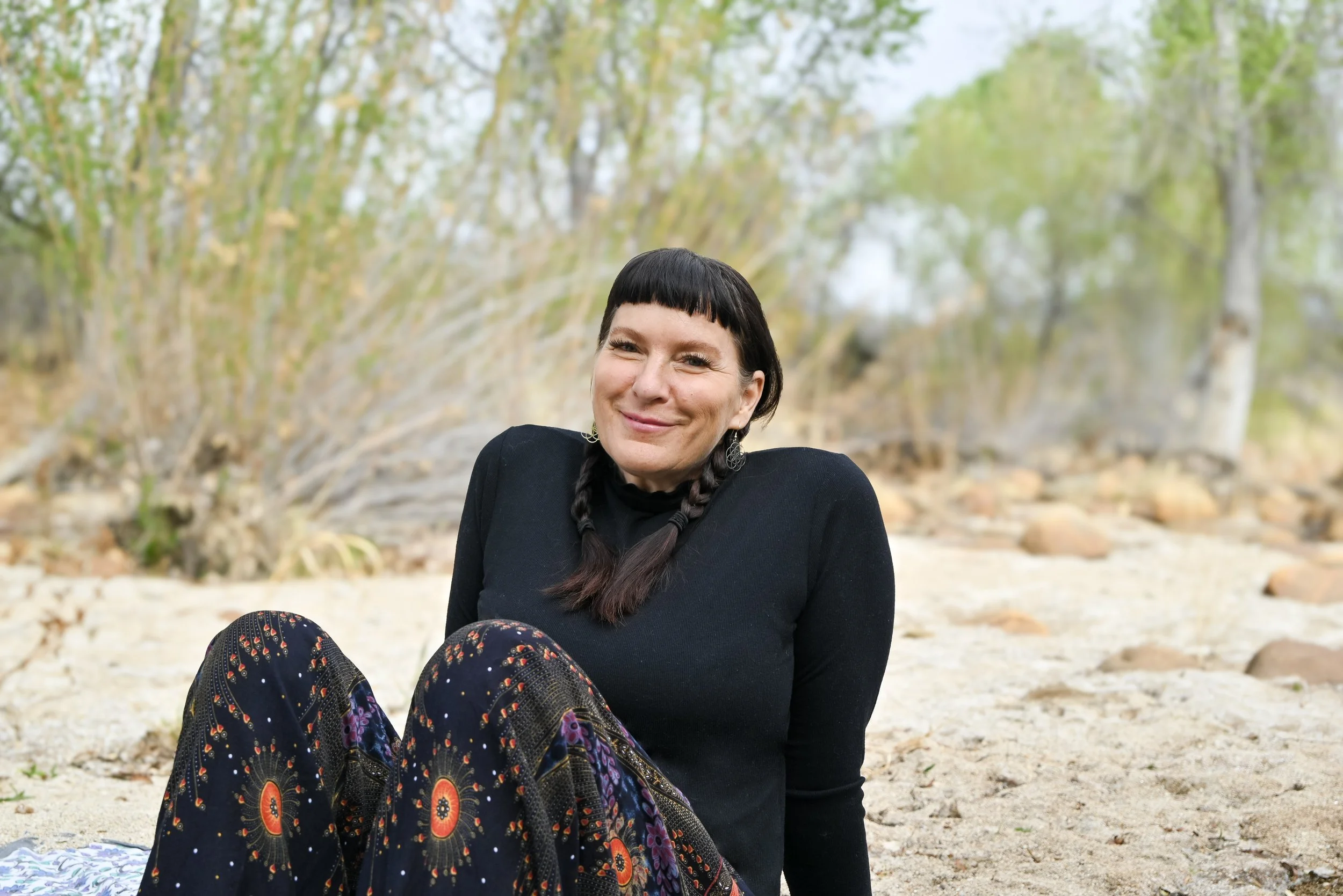 A woman with short dark hair and braids, smiling and sitting on a sandy beach with trees in the background.