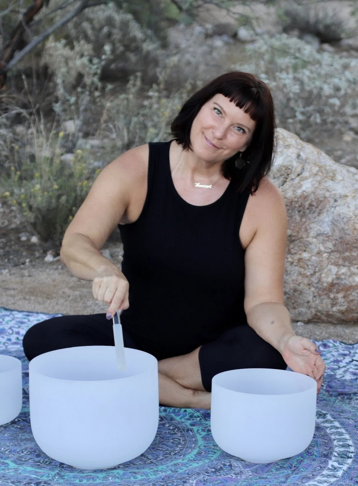 A woman with dark hair and a black sleeveless top sitting on a blanket outdoors, playing singing bowls.