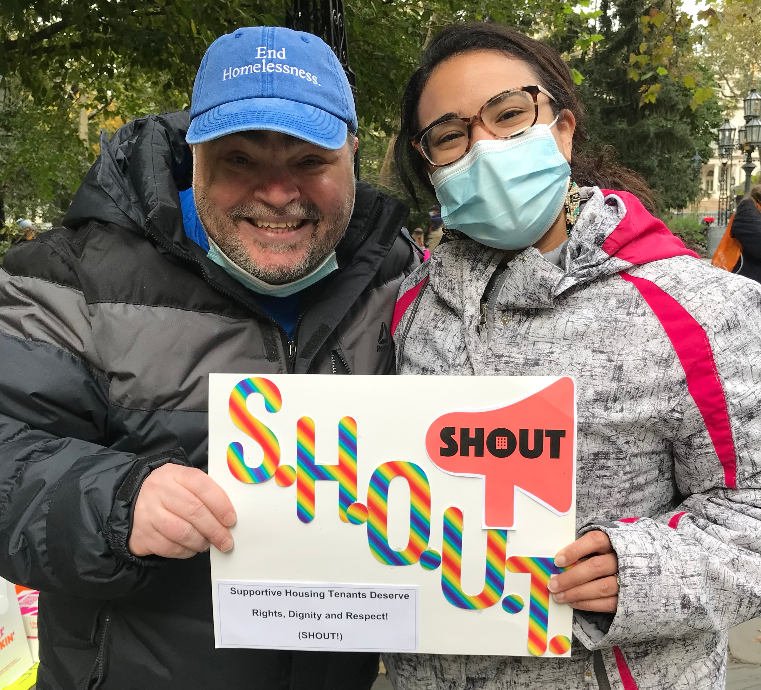 Two tenants organizers standing outside and holding a sign with the SHOUT logo and a caption on the sign that reads "Support Housing Tenants Deserve Rights, Dignity, and Respect! (SHOUT!)