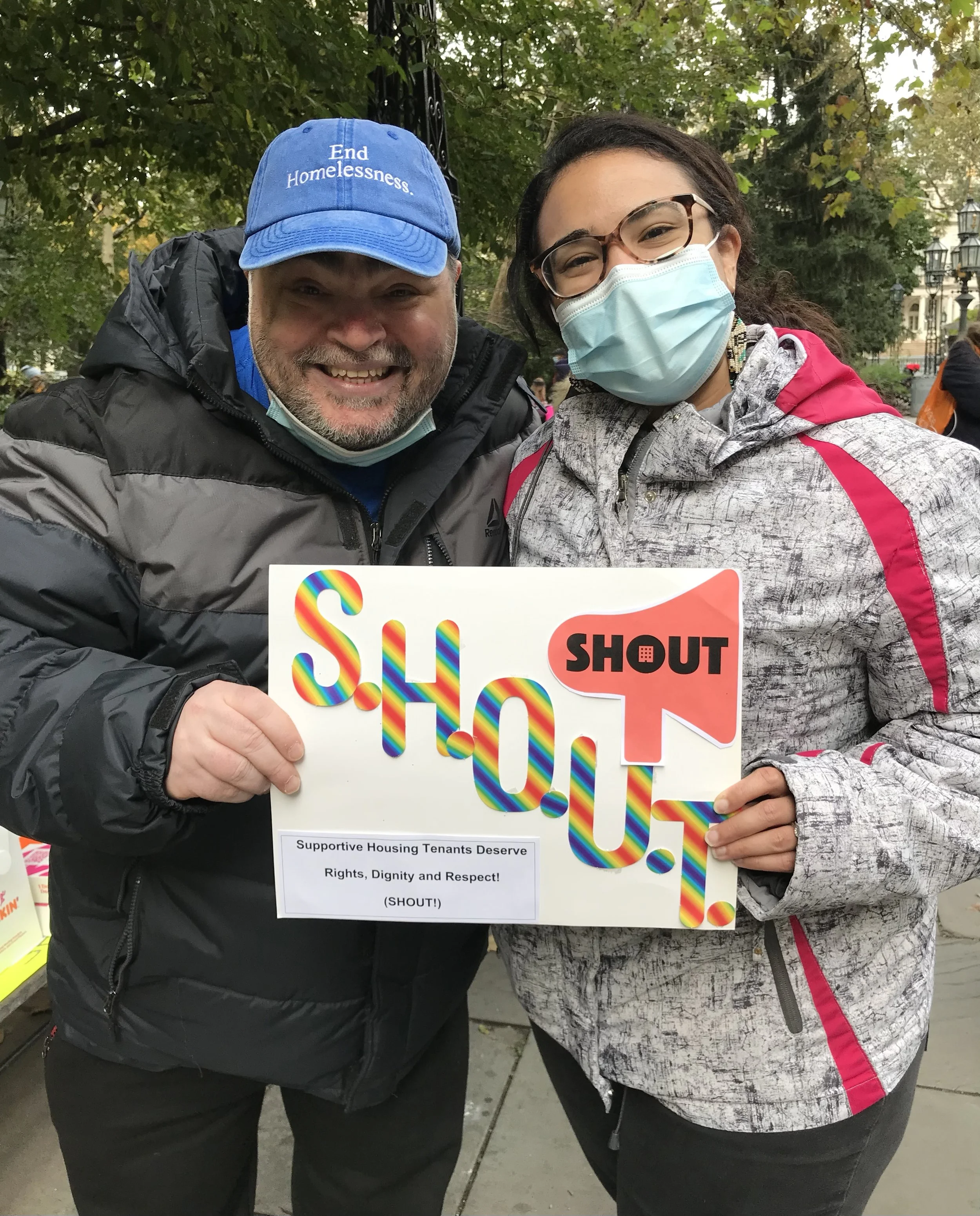 Two tenants organizers standing outside and holding a sign with the SHOUT logo and a caption on the sign that reads "Support Housing Tenants Deserve Rights, Dignity, and Respect! (SHOUT!)