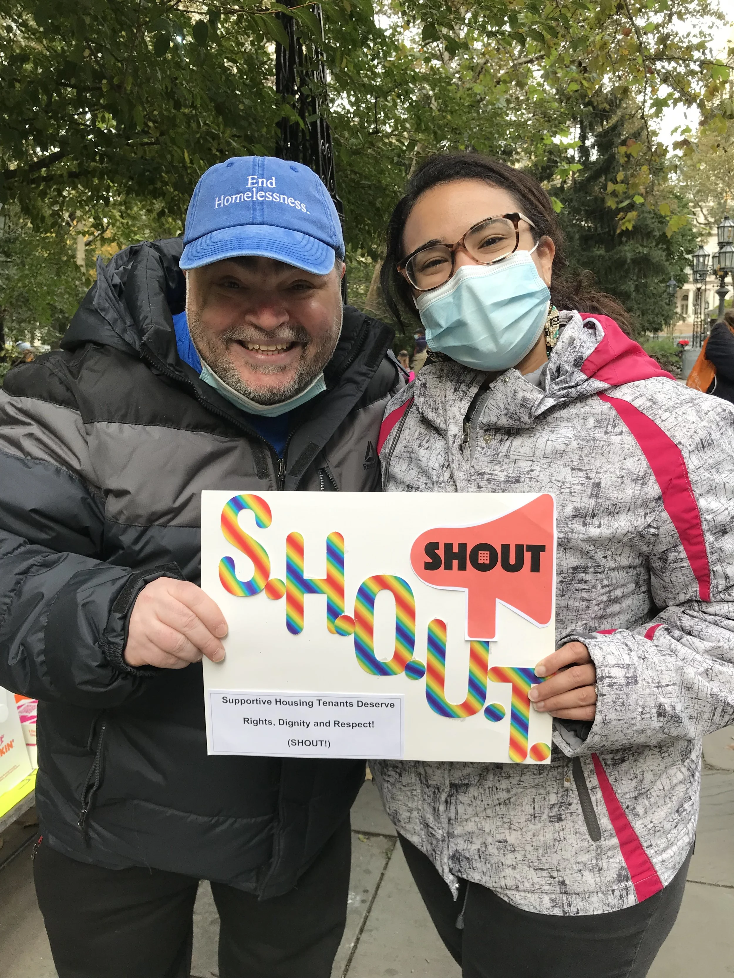 Two tenants organizers standing outside and holding a sign with the SHOUT logo and a caption on the sign that reads "Support Housing Tenants Deserve Rights, Dignity, and Respect! (SHOUT!)