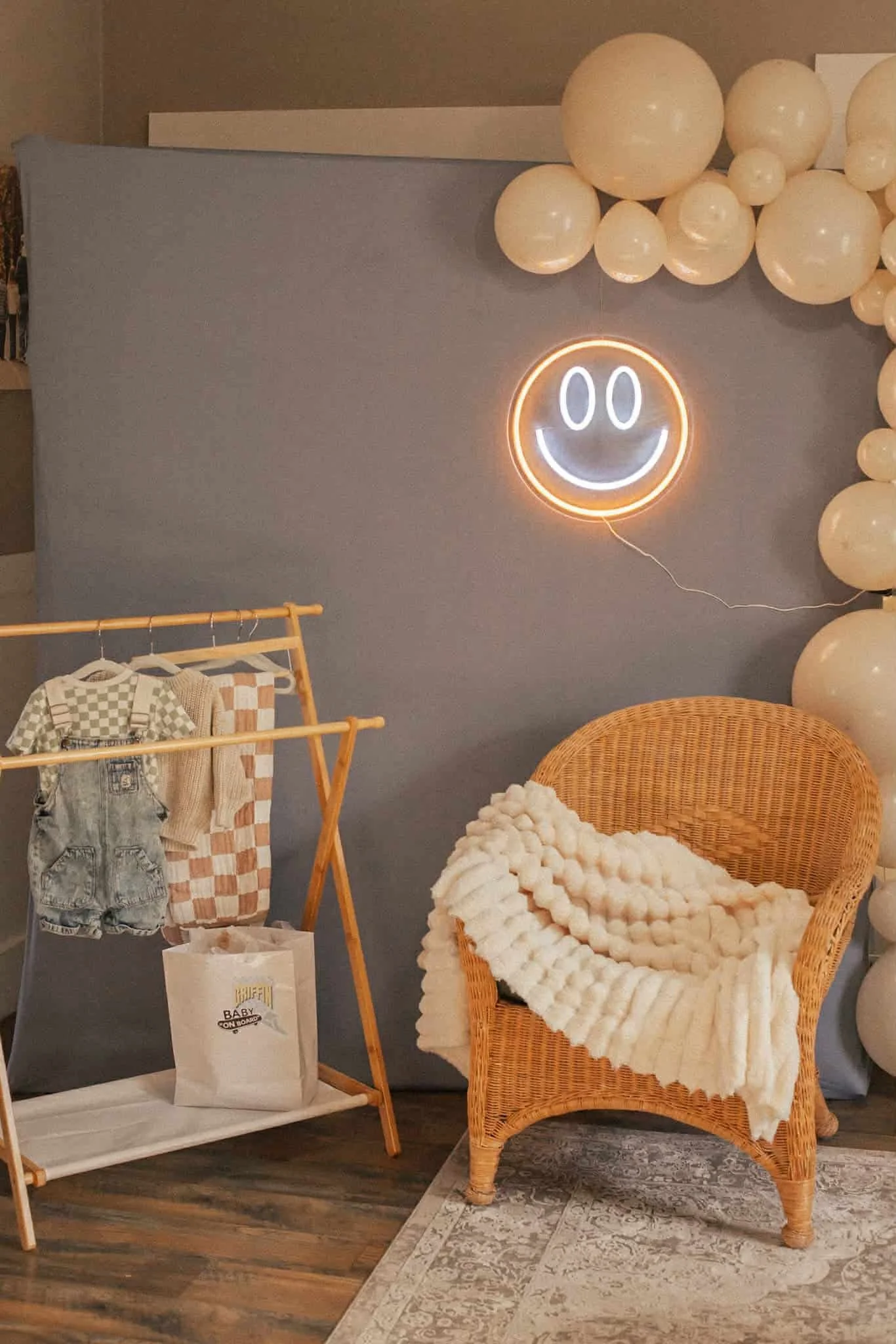 Baby shower photo backdrop for boy Vans theme. Wicker chair and plush white blanket, blue backdrop with neon smiley face sign and beige balloons. A clothing rack with checkered baby clothes and blanket.