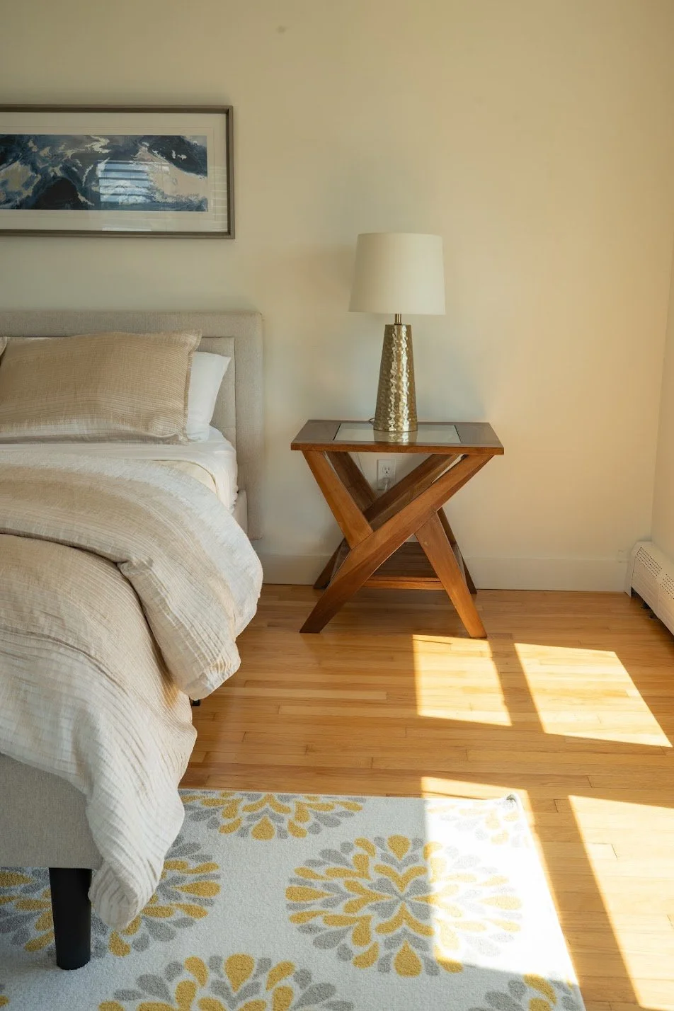 A bedroom corner with a bed, beige bedding, a wooden nightstand, a table lamp with a textured gold base, a framed artwork hanging above, and sunlight streaming onto a patterned rug and hardwood floor.