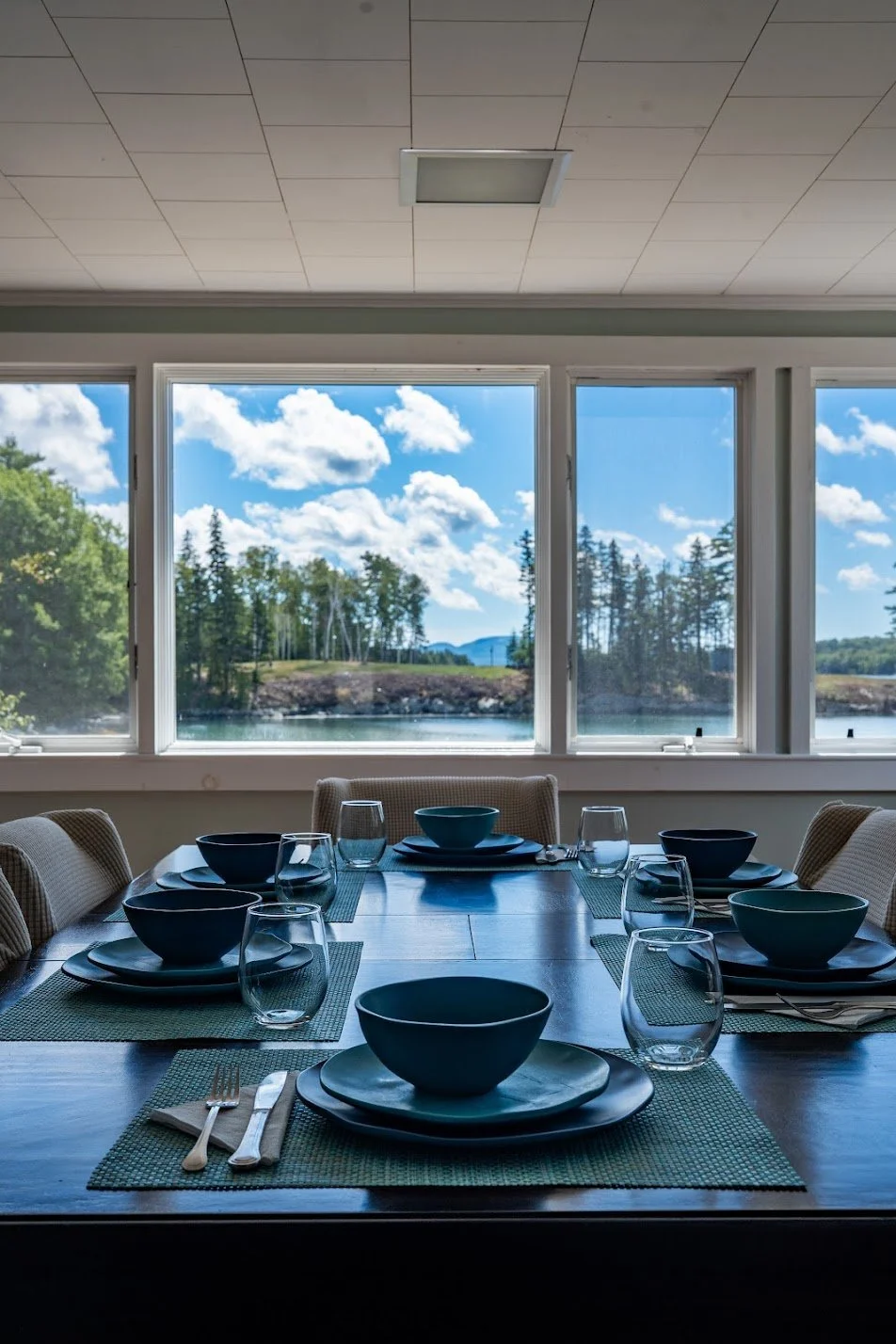 Dining table set with bowls, glasses, silverware, and placemats, with large windows showing a lakeside view of trees, water, and a blue sky with clouds inside a well-lit room.