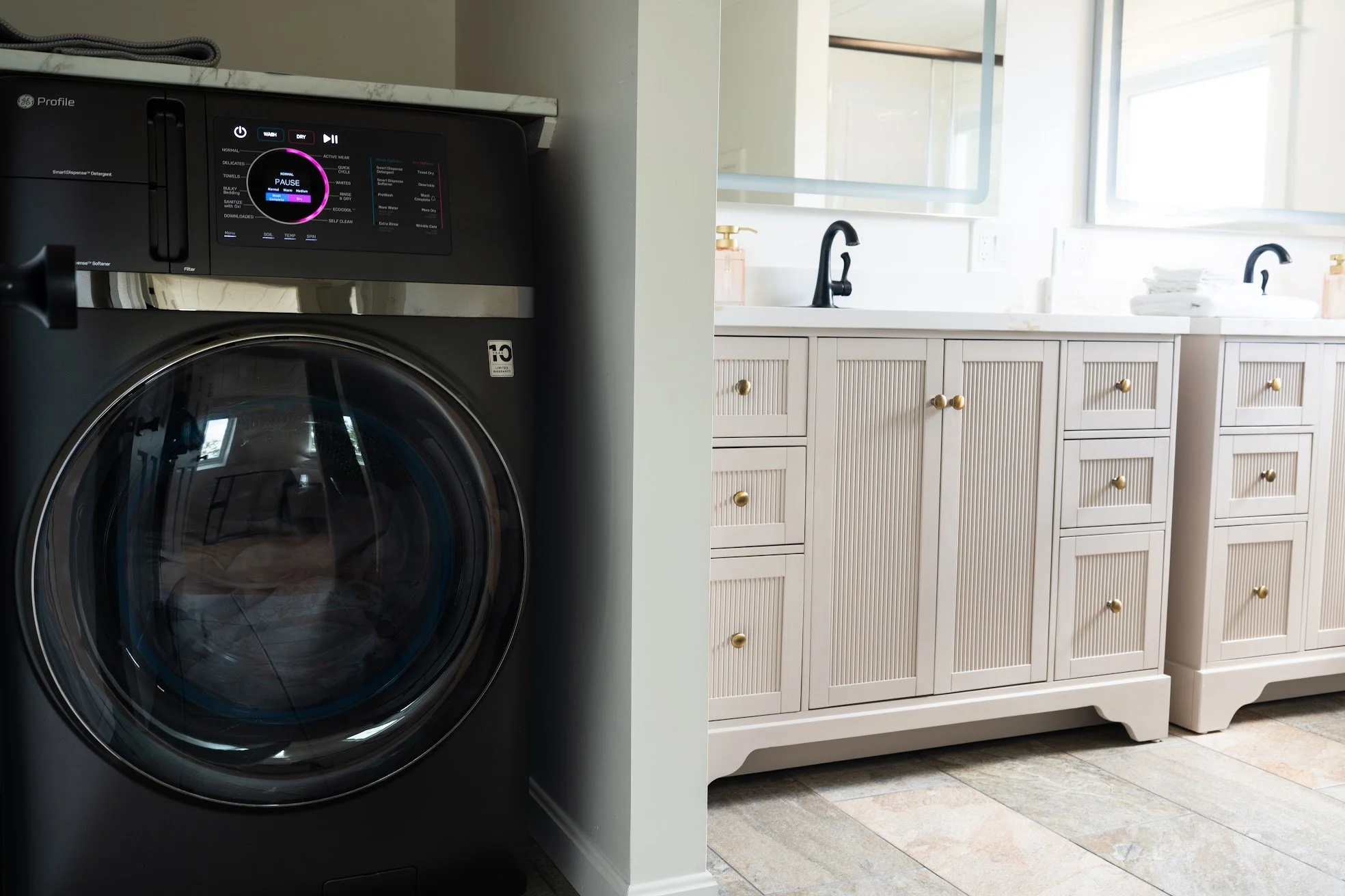 Laundry room with a front-loading washing machine on the left and beige cabinets with black sinks and gold knobs on the right, illuminated by natural light from windows.