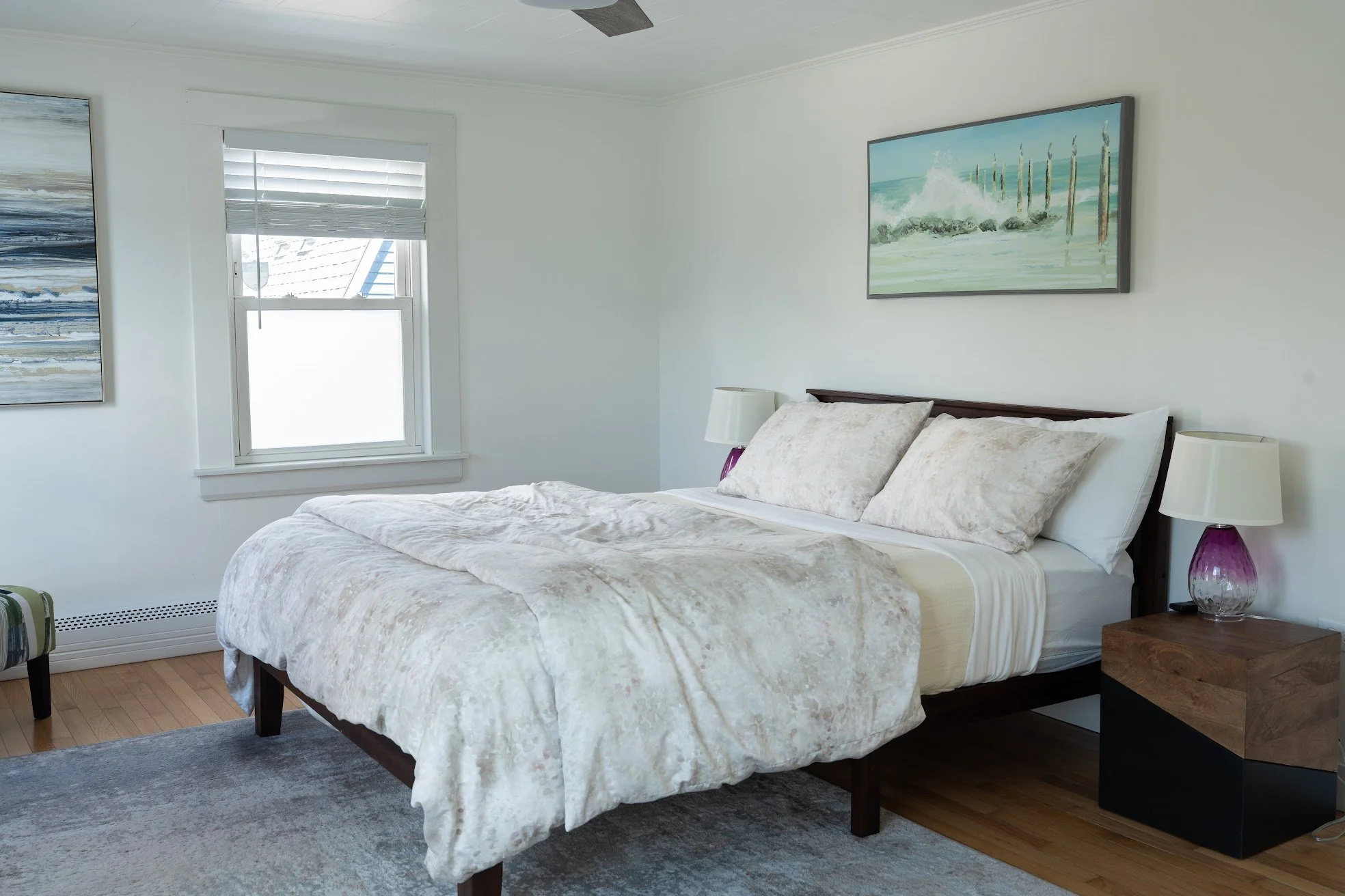 A bedroom with a bed, white bedding, pillows, two nightstands with lamps, a window, and wall art.