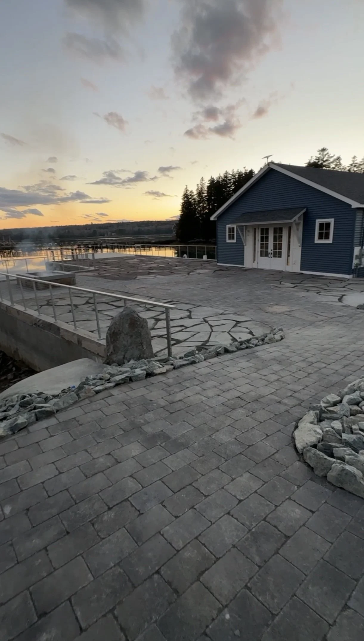 A scenic view of a blue house with white doors and windows near a body of water at sunset. The foreground features a paved area with stone and brick patterns, with some rock borders and a metal railing.