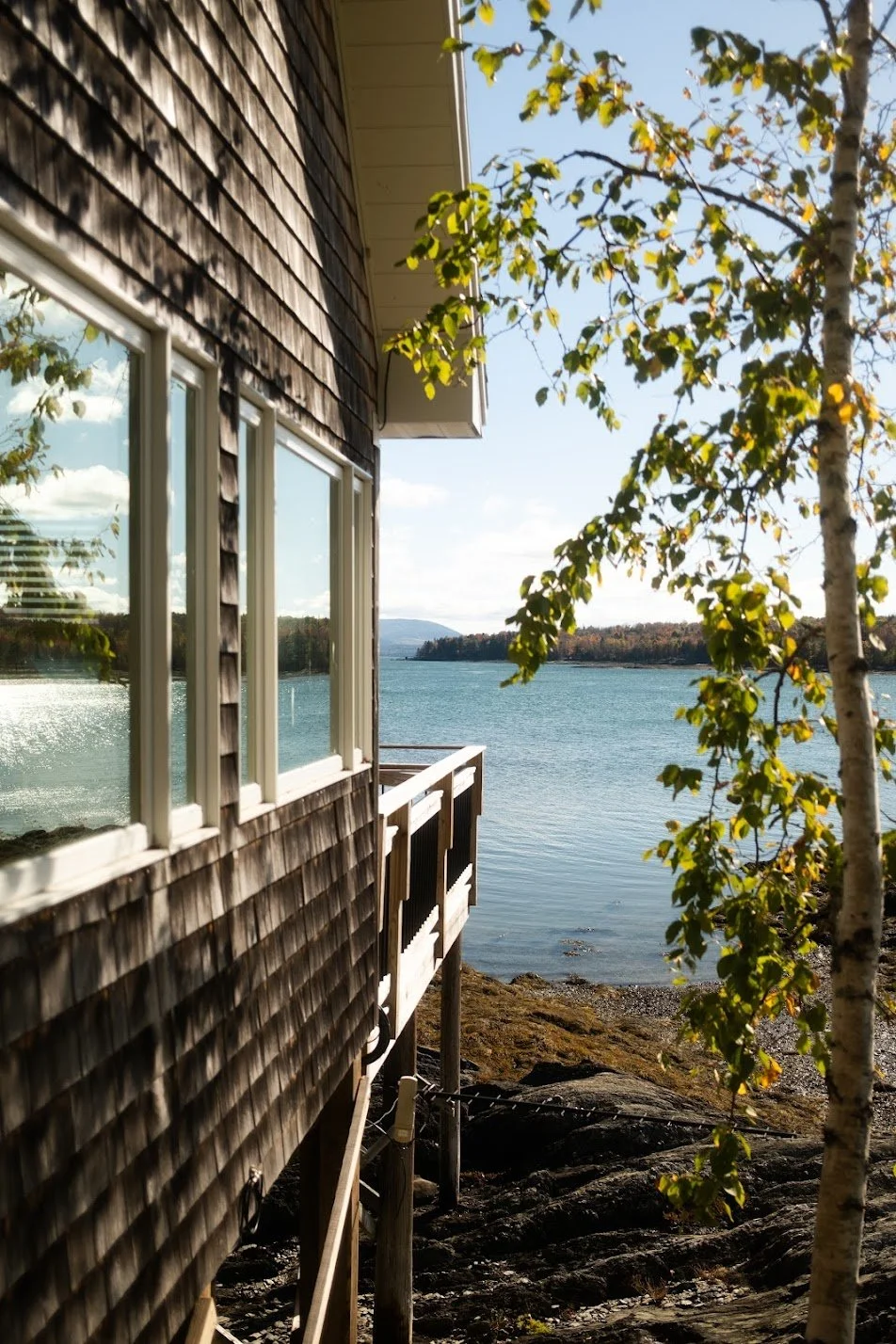 View of a lakeside house with wooden siding, multiple windows reflecting the sky, and a small balcony overlooking the water, framed by a tree with green leaves.