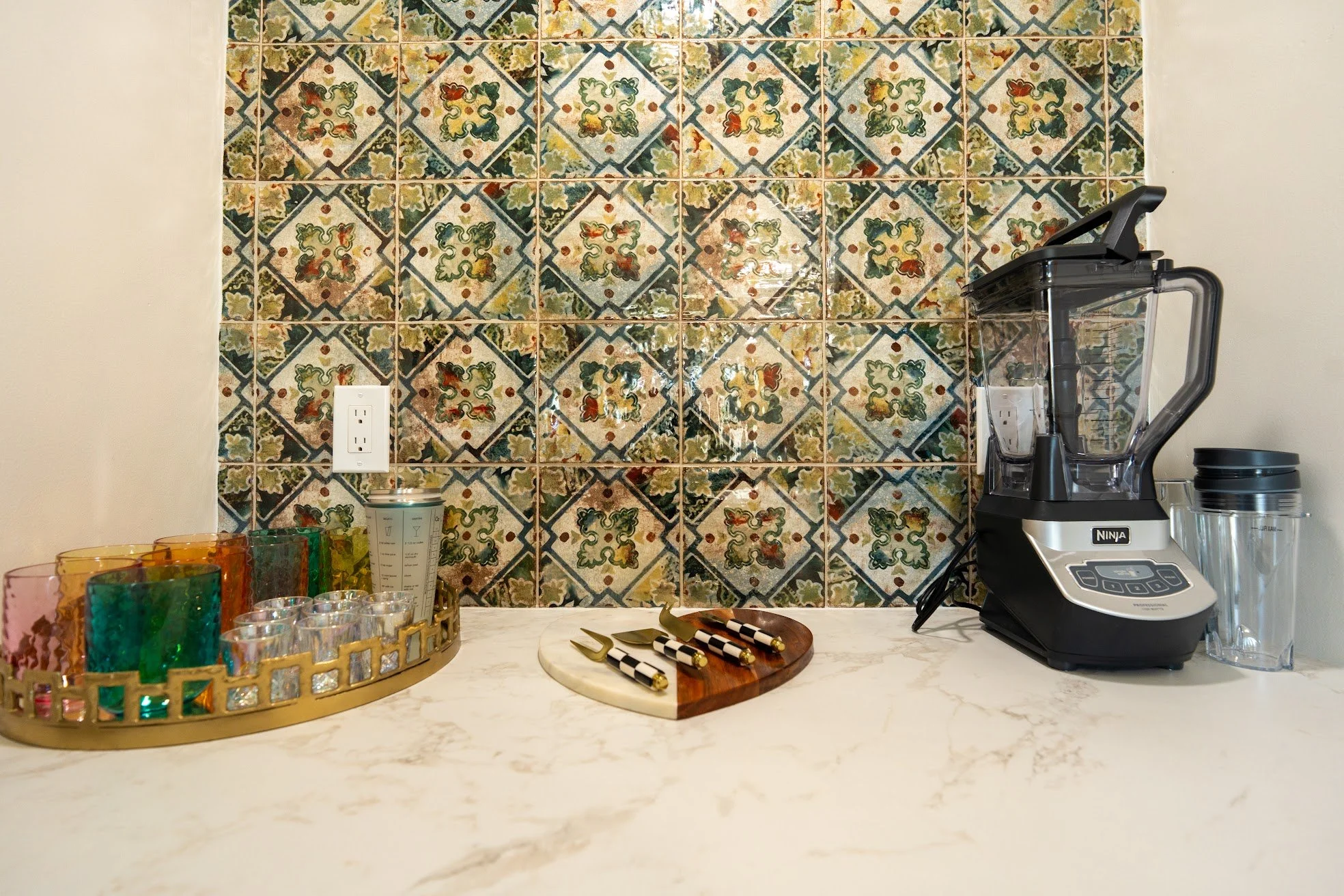 Kitchen countertop with colorful glassware tray, black and white checkered knives on a wooden and marble cutting board, Ninja blender, and a wall with floral-patterned tiles.